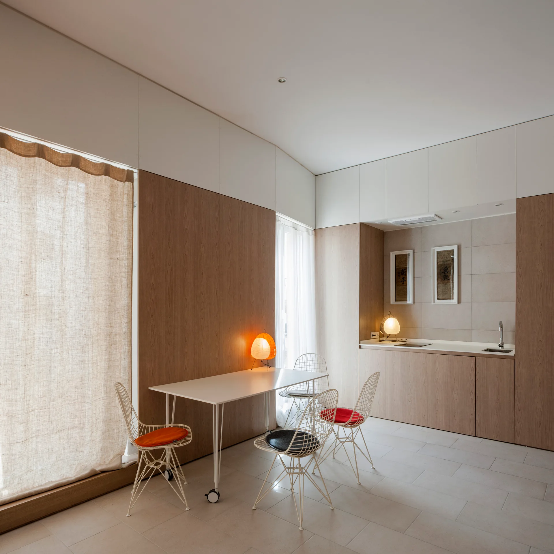 A minimalist dining area featuring a white table with wire chairs next to a wood-paneled wall and integrated kitchenette.