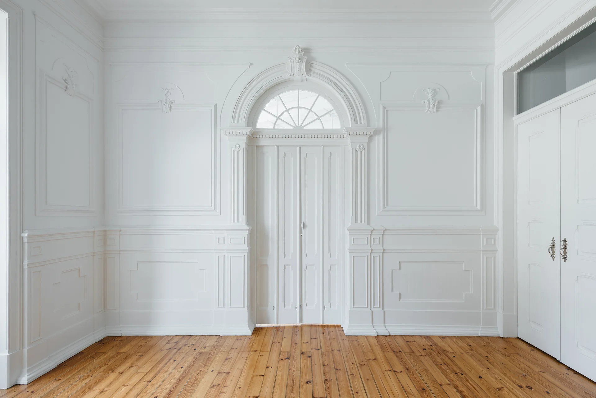 An empty white room features ornate wall molding, wainscoting, a tall white door with an arched transom window, and natural pine wood flooring.