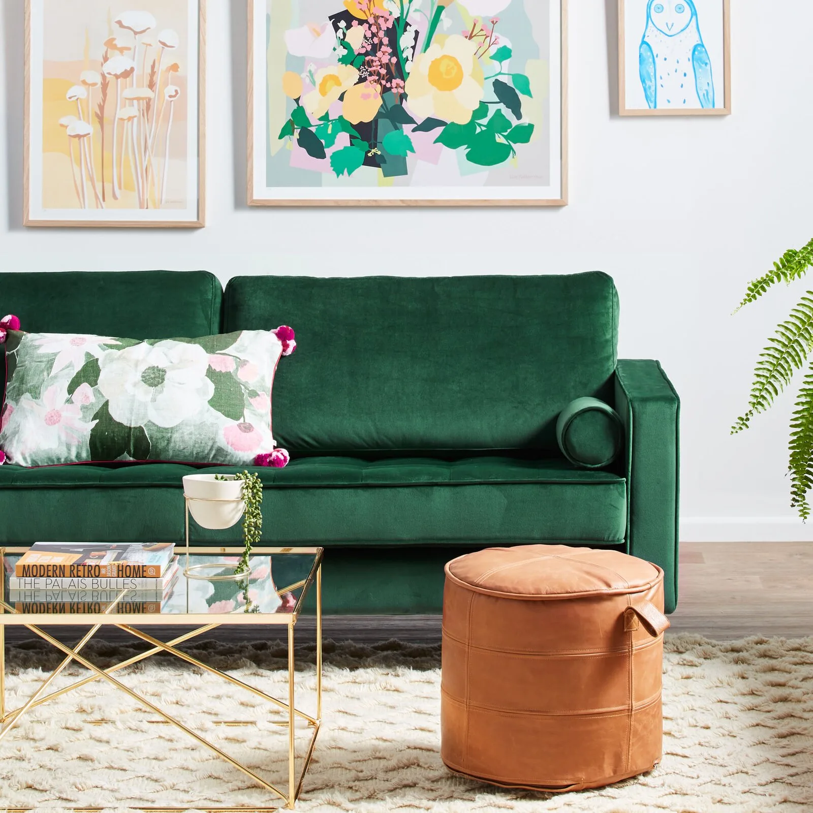 A green velvet sofa with a floral pillow is paired with a tan leather ottoman and gold glass coffee table on a shag rug.