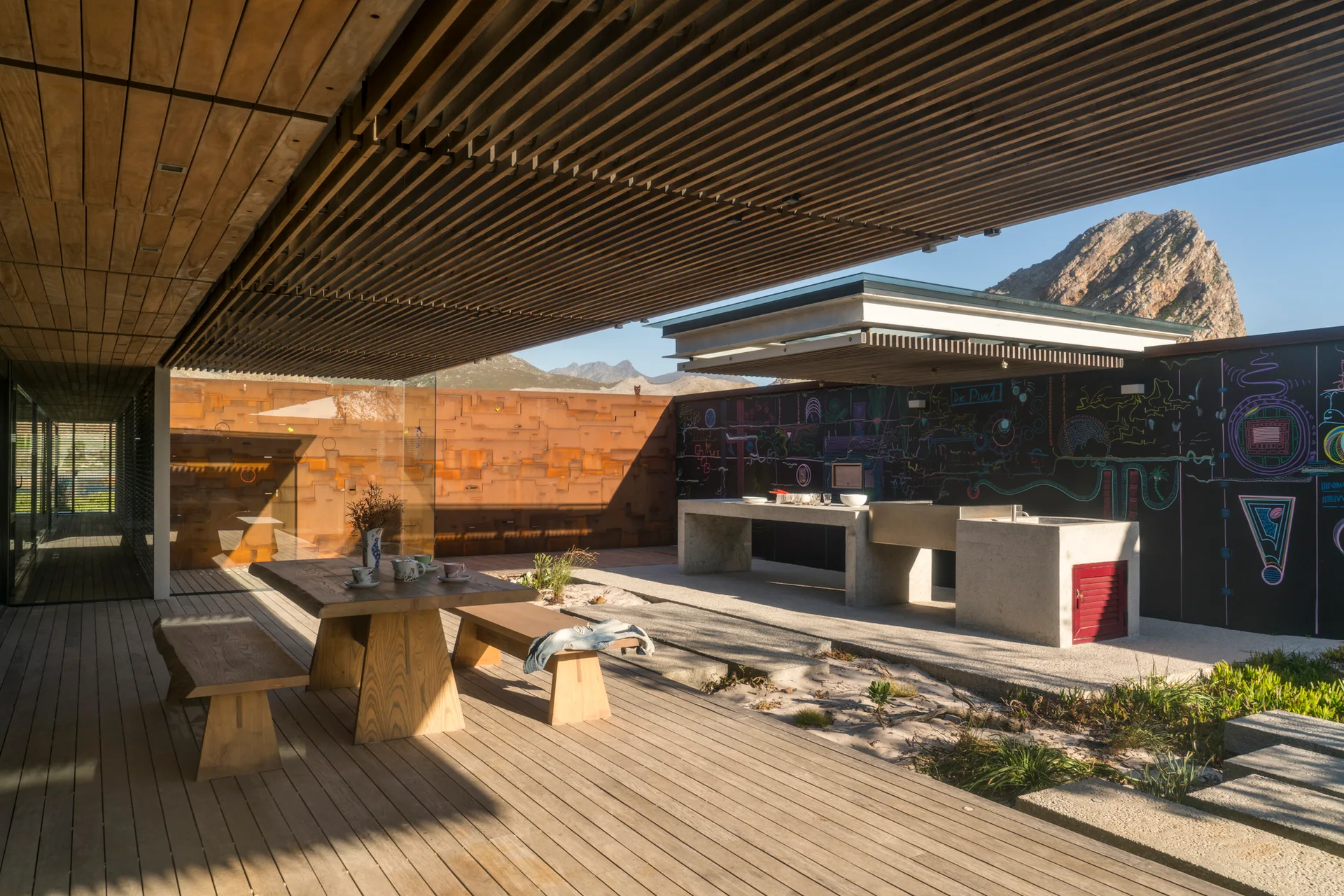 A modern covered patio with a wooden dining table, a concrete outdoor kitchen counter, a weathered metal wall, and a colorful chalkboard.