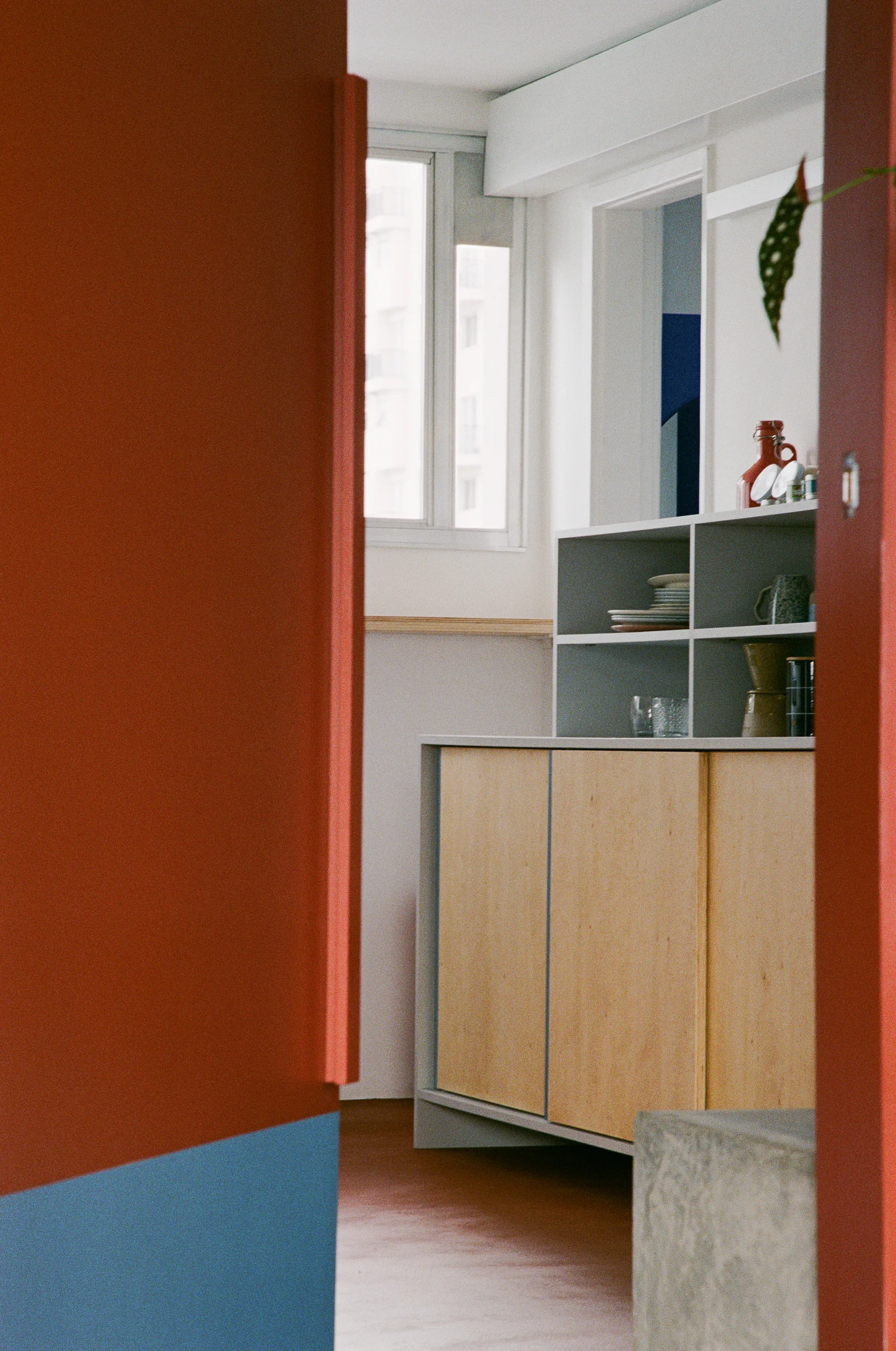 A view through a bold red and blue doorway reveals a light wood kitchen cabinet with grey open shelving displaying ceramic plates and glasses.