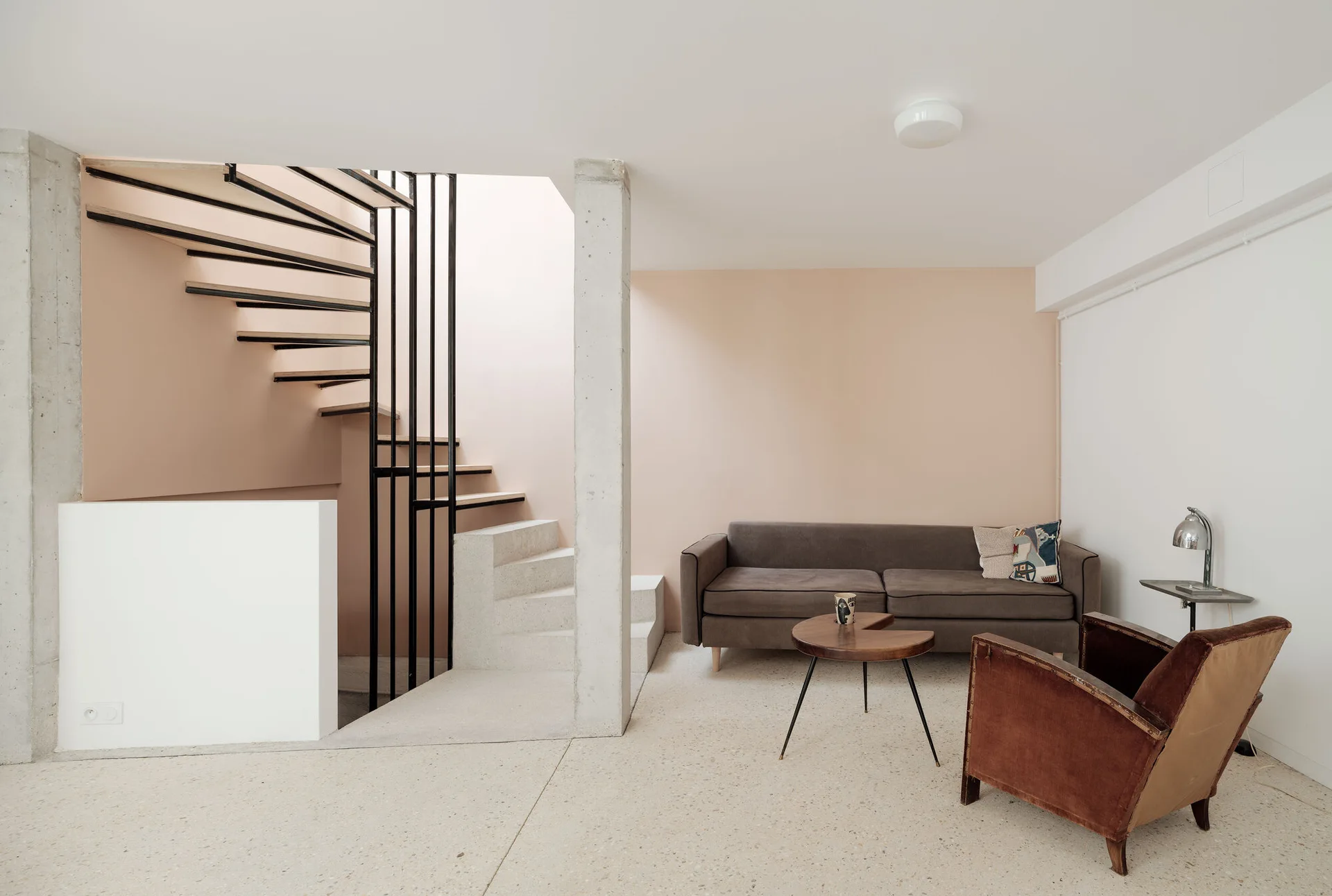 A minimalist living room featuring a grey sofa against a blush pink wall, a brown armchair, and an open-riser spiral staircase by a concrete column.