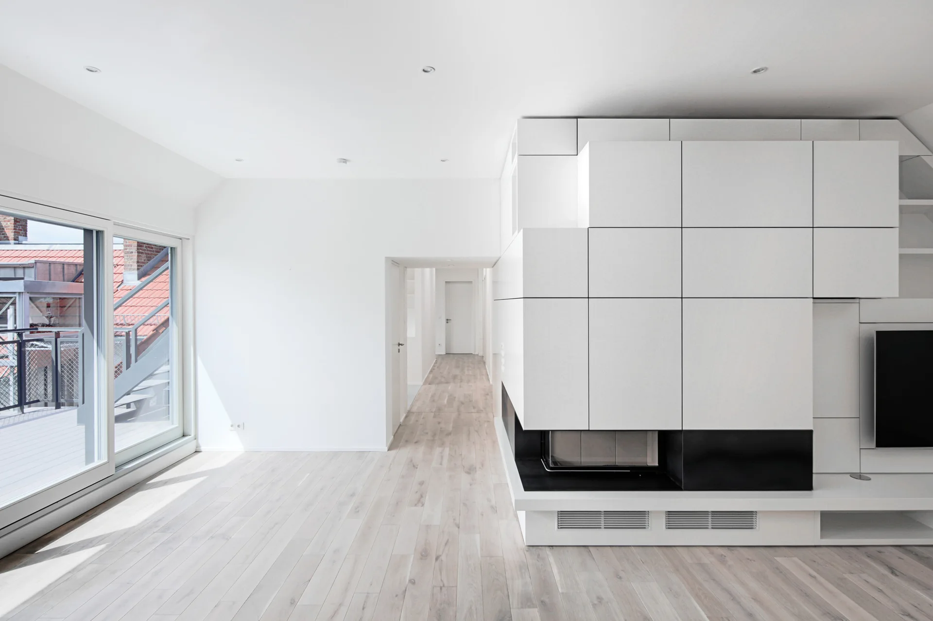 Minimalist living space with a blocky white modular fireplace wall, light wood plank floors, and a large sliding glass door opening to a balcony.