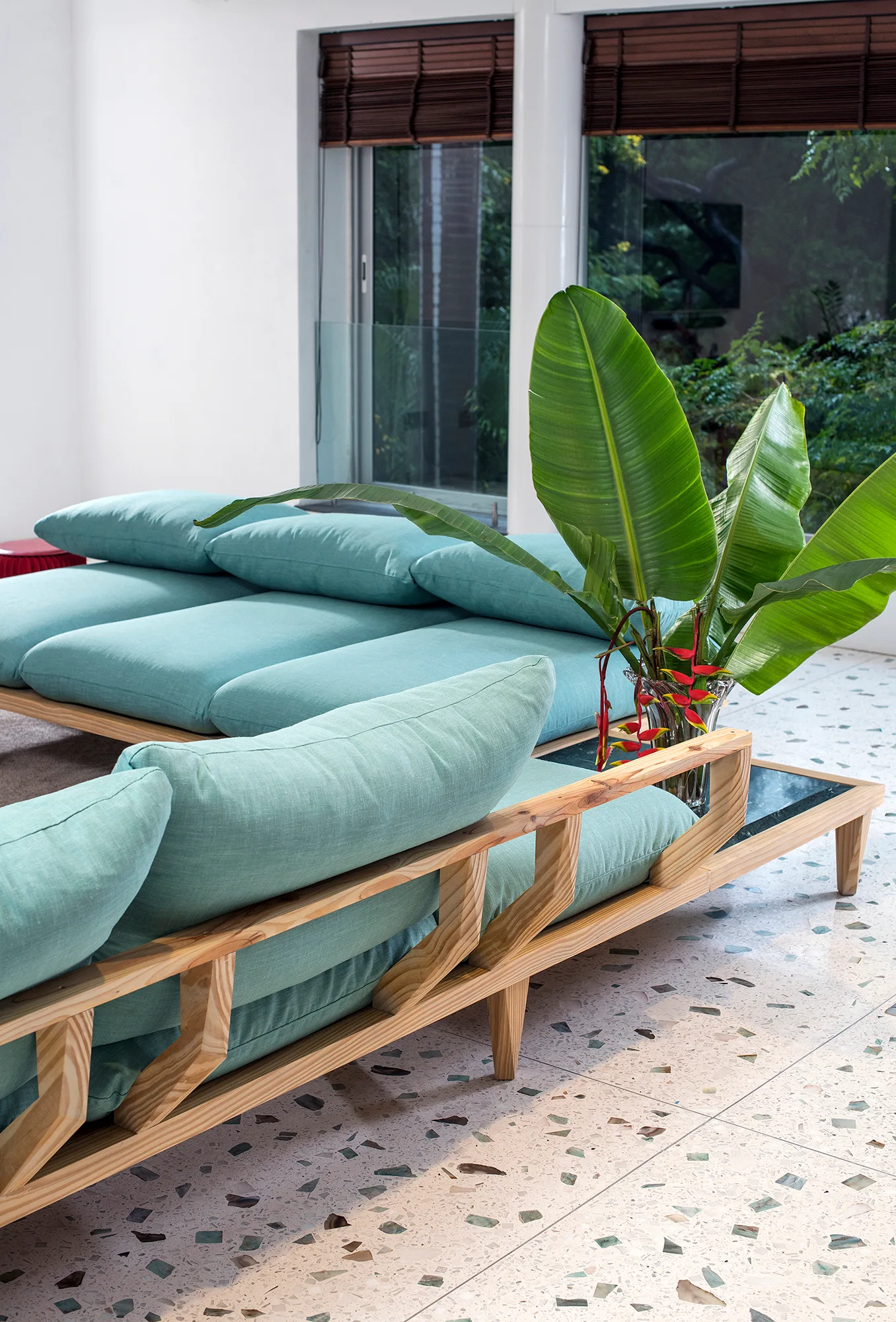 A modern low-profile teal sofa with an exposed light wood frame sits on speckled terrazzo tile flooring near a large tropical plant.