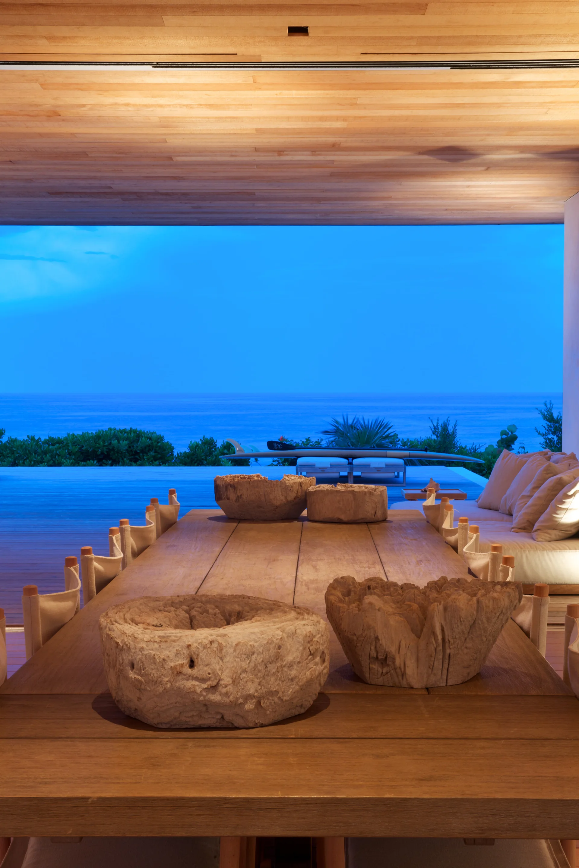 A covered wooden patio features a long dining table set with rustic wooden bowls, canvas chairs, and an infinity pool overlooking the ocean at dusk.