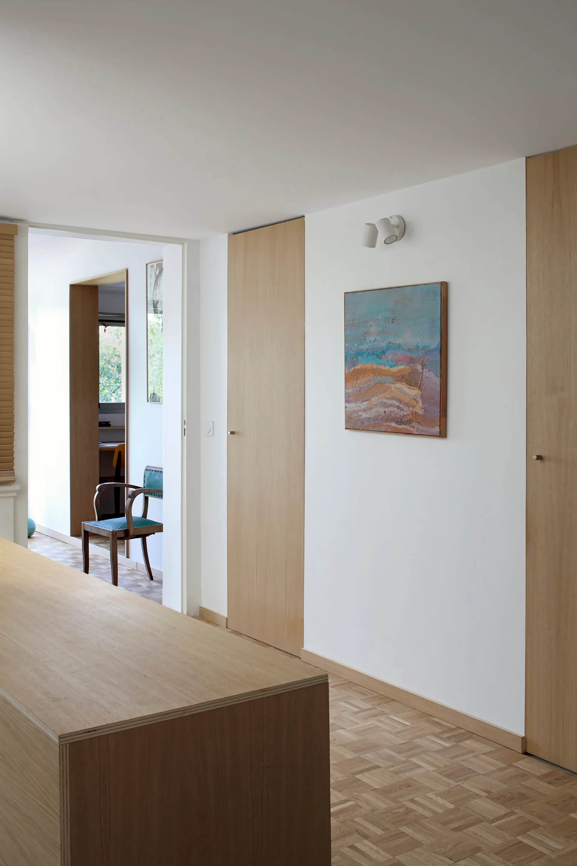 A minimalist white hallway featuring full-height light wood doors, light oak parquet flooring, and an abstract painting illuminated by a spotlight.
