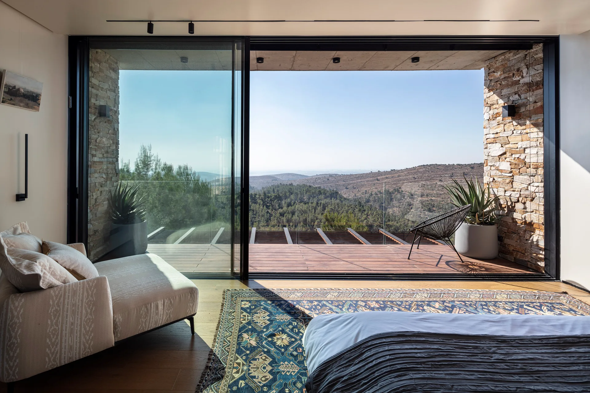 A modern bedroom features an armchair and patterned rug next to large sliding glass doors that open to a wooden deck with stone walls and a scenic view.
