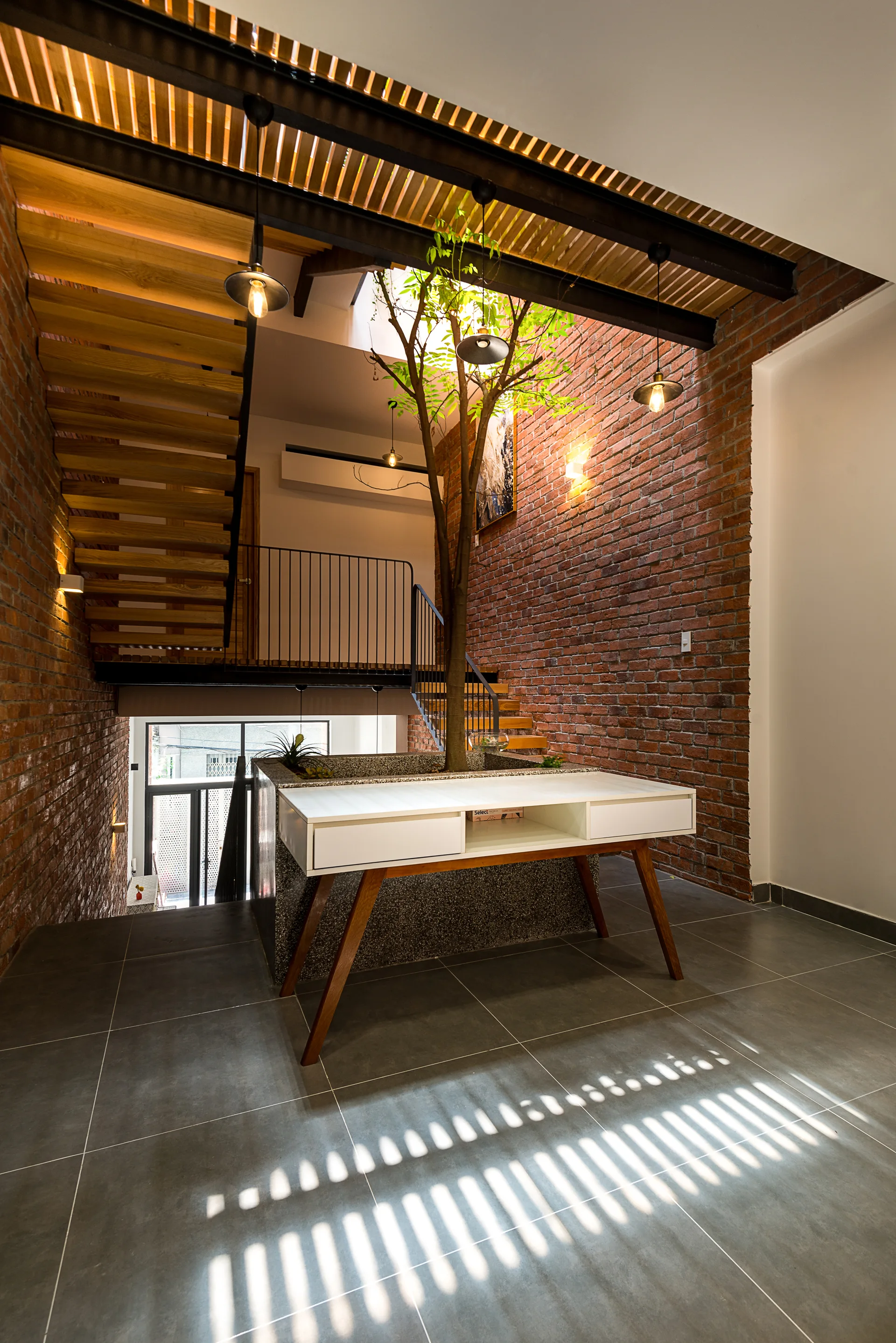 An indoor tree grows from a stone planter beside a wooden staircase, flanked by exposed red brick walls and a slatted timber ceiling.