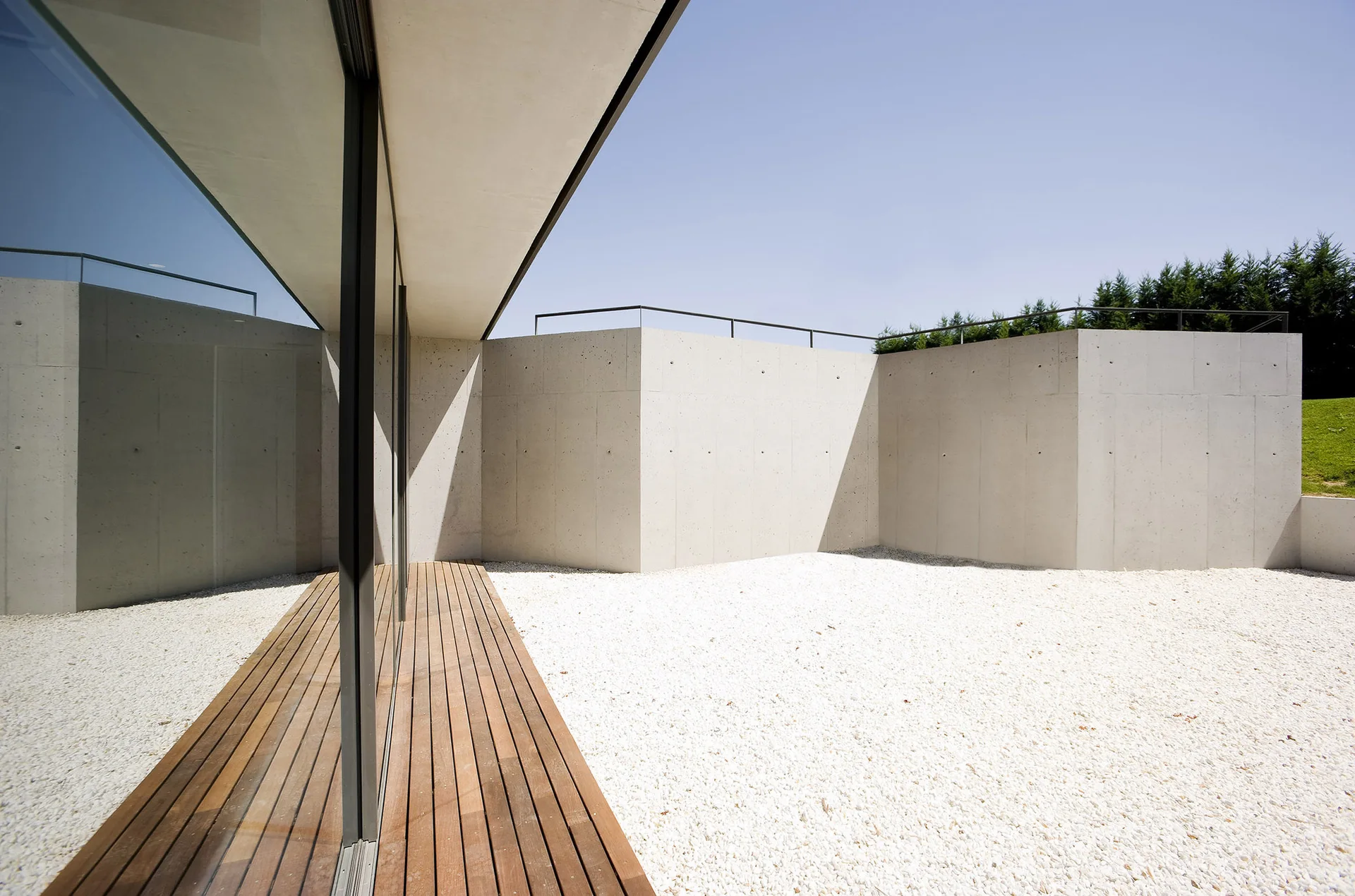 A modern outdoor courtyard featuring light grey concrete walls, a slatted wooden deck pathway, and white gravel ground cover under a blue sky.