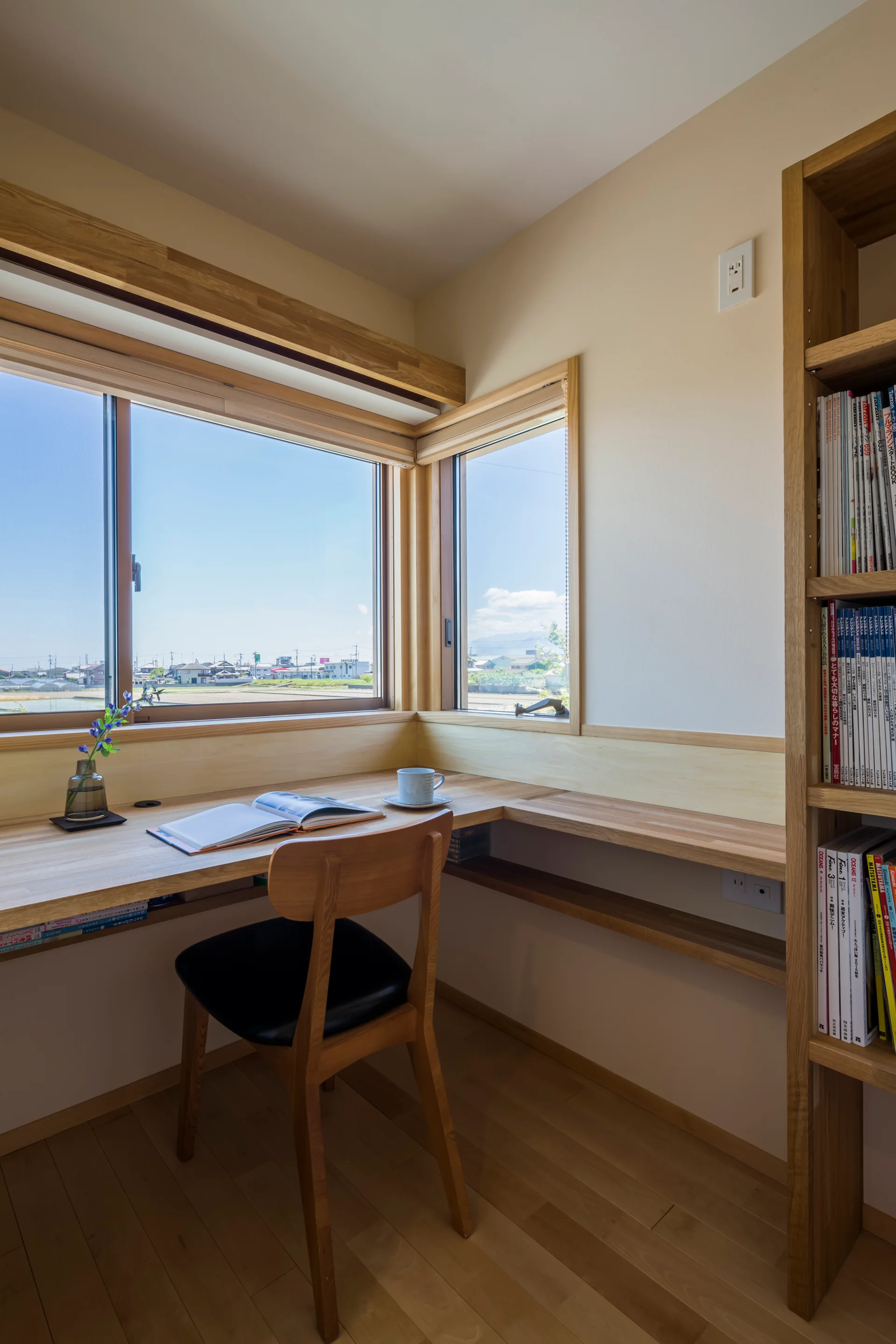 A minimalist home office features an L-shaped built-in wooden desk under a corner window, with a wooden chair and a tall matching bookshelf.