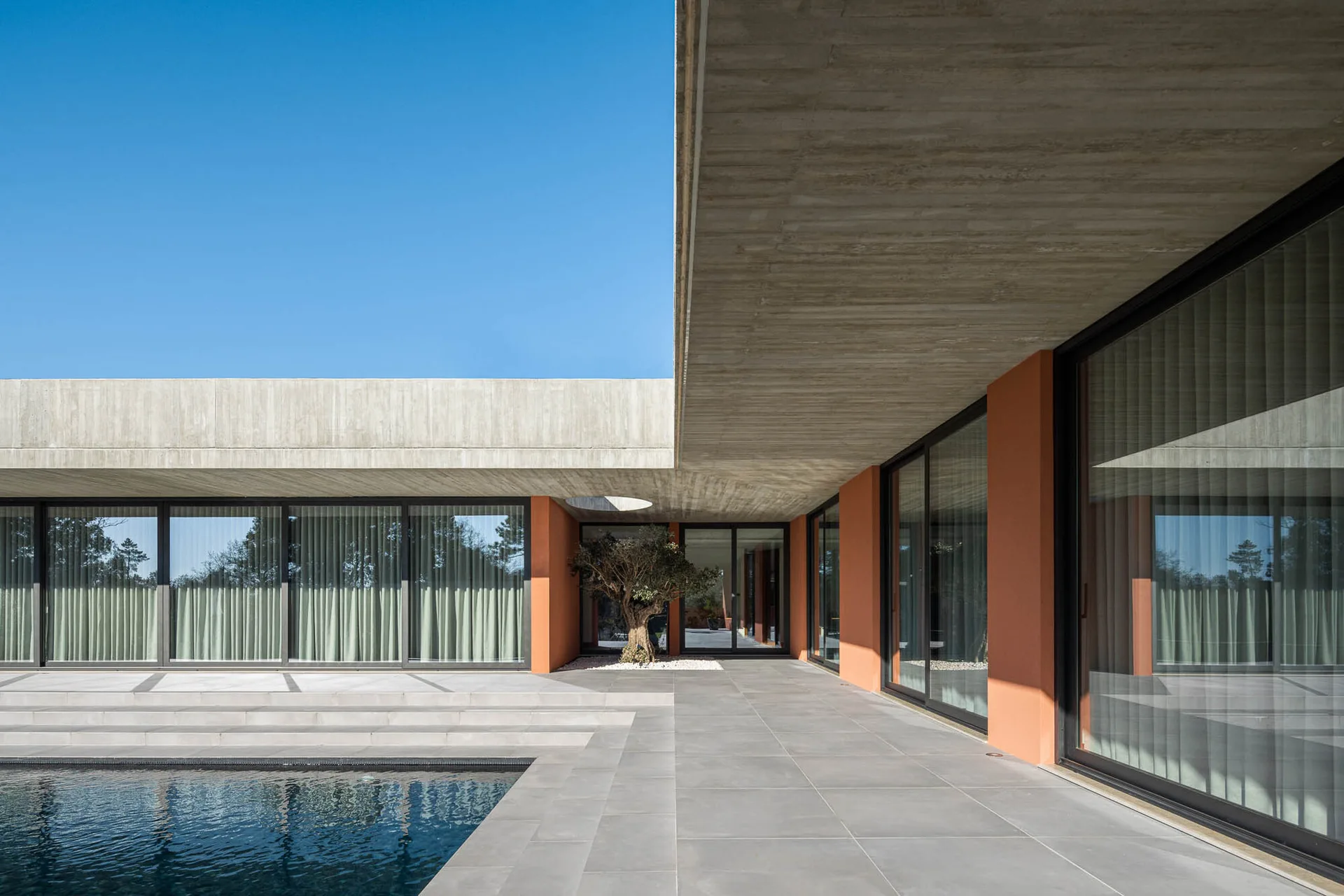 A modernist outdoor pool patio features a grey tiled deck, terracotta-colored walls, glass sliding doors, and a raw concrete roof overhang under a blue sky.