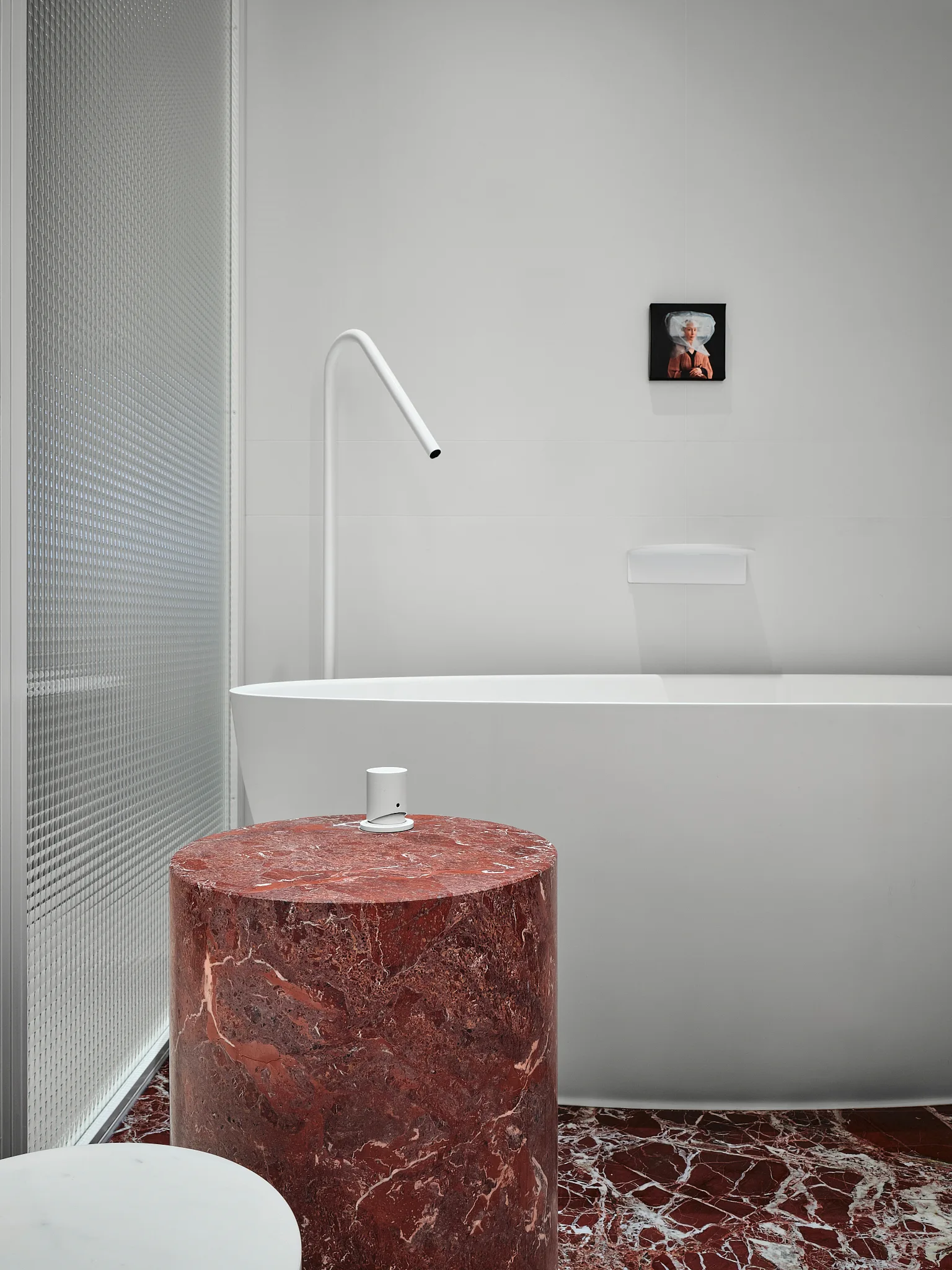 A minimalist bathroom features a white freestanding tub, a red veined marble floor, a matching cylindrical pedestal table, and a textured glass partition.