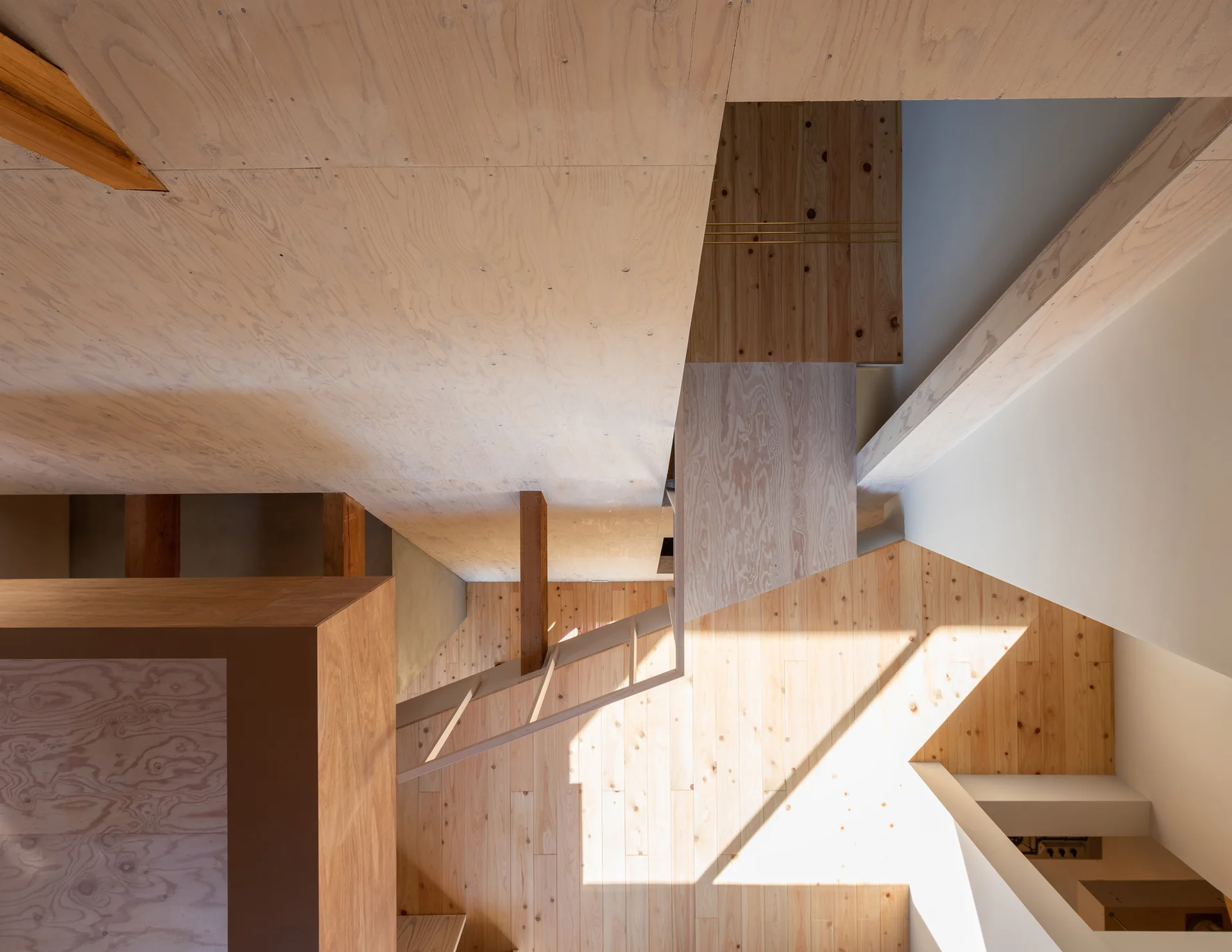 A top-down view of a minimalist stairwell with light plywood paneled walls, exposed wood beams, and natural sunlight casting geometric shadows.