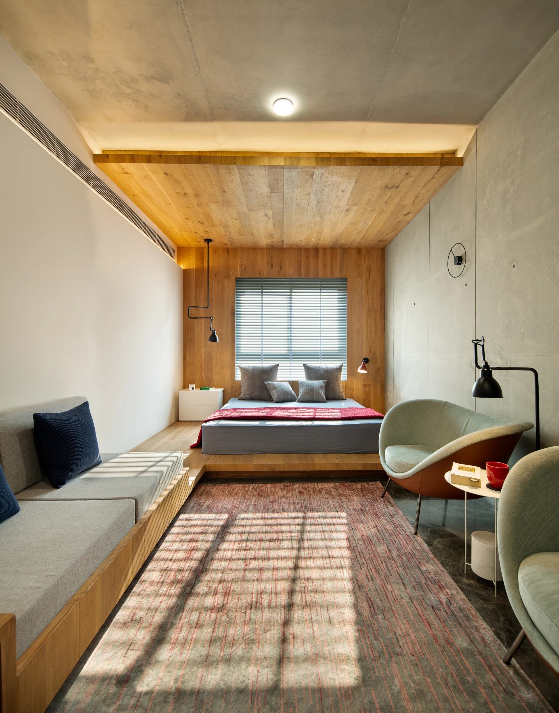 A modern bedroom features a platform bed framed by warm wood paneling, a raw concrete wall, a grey built-in sofa, and two scoop-shaped armchairs.