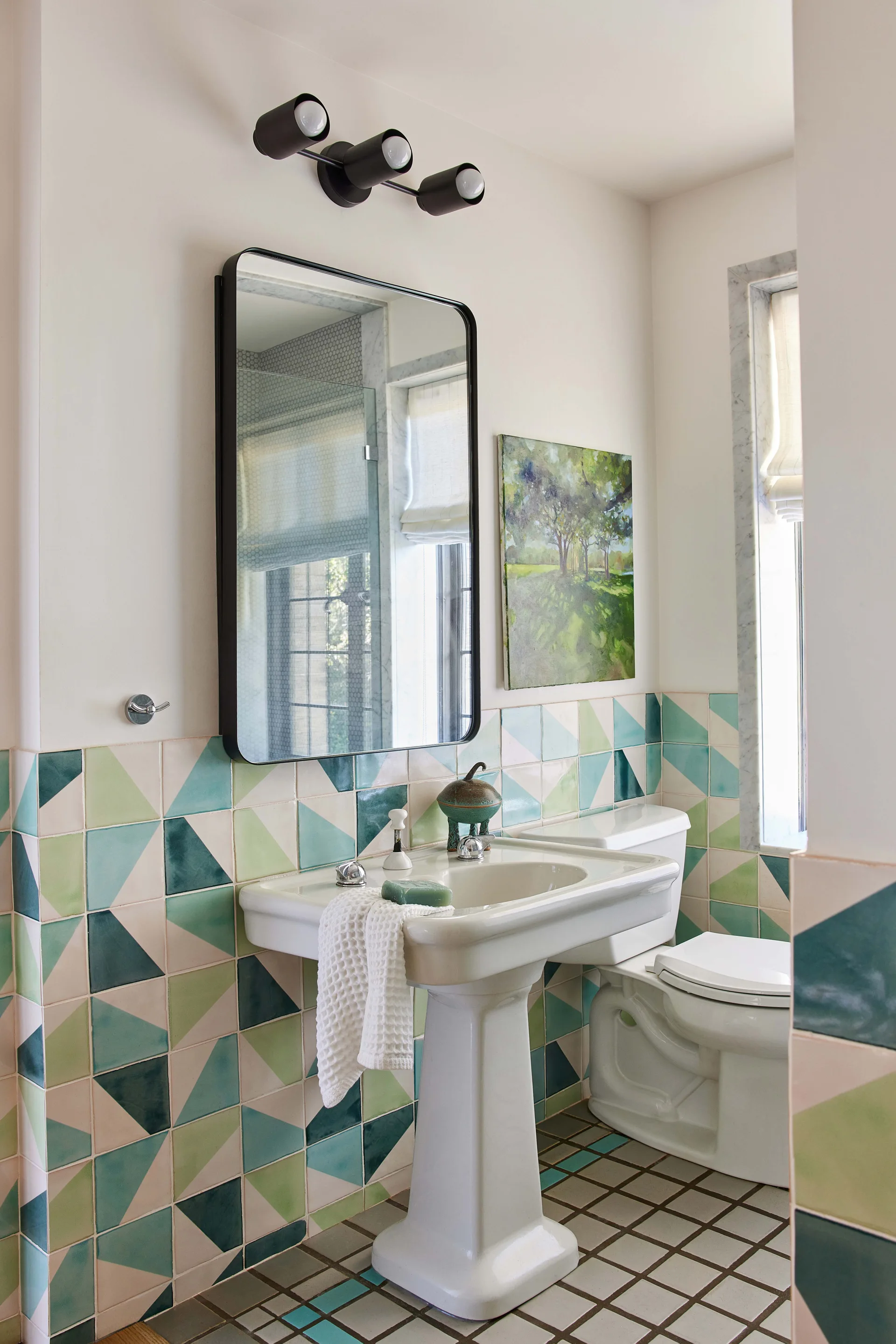 A bathroom featuring a white pedestal sink, a black-framed mirror, and lower walls decorated with green, blue, and cream geometric triangular tiles.