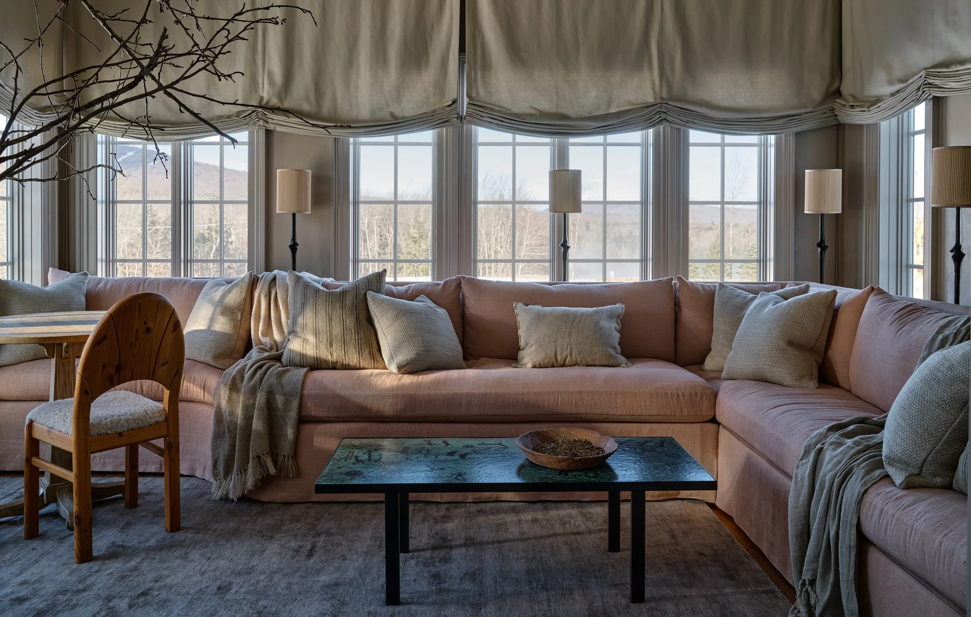 A large blush pink sectional sofa sits in front of multi-pane windows with beige valances, alongside a low coffee table and wooden chair.