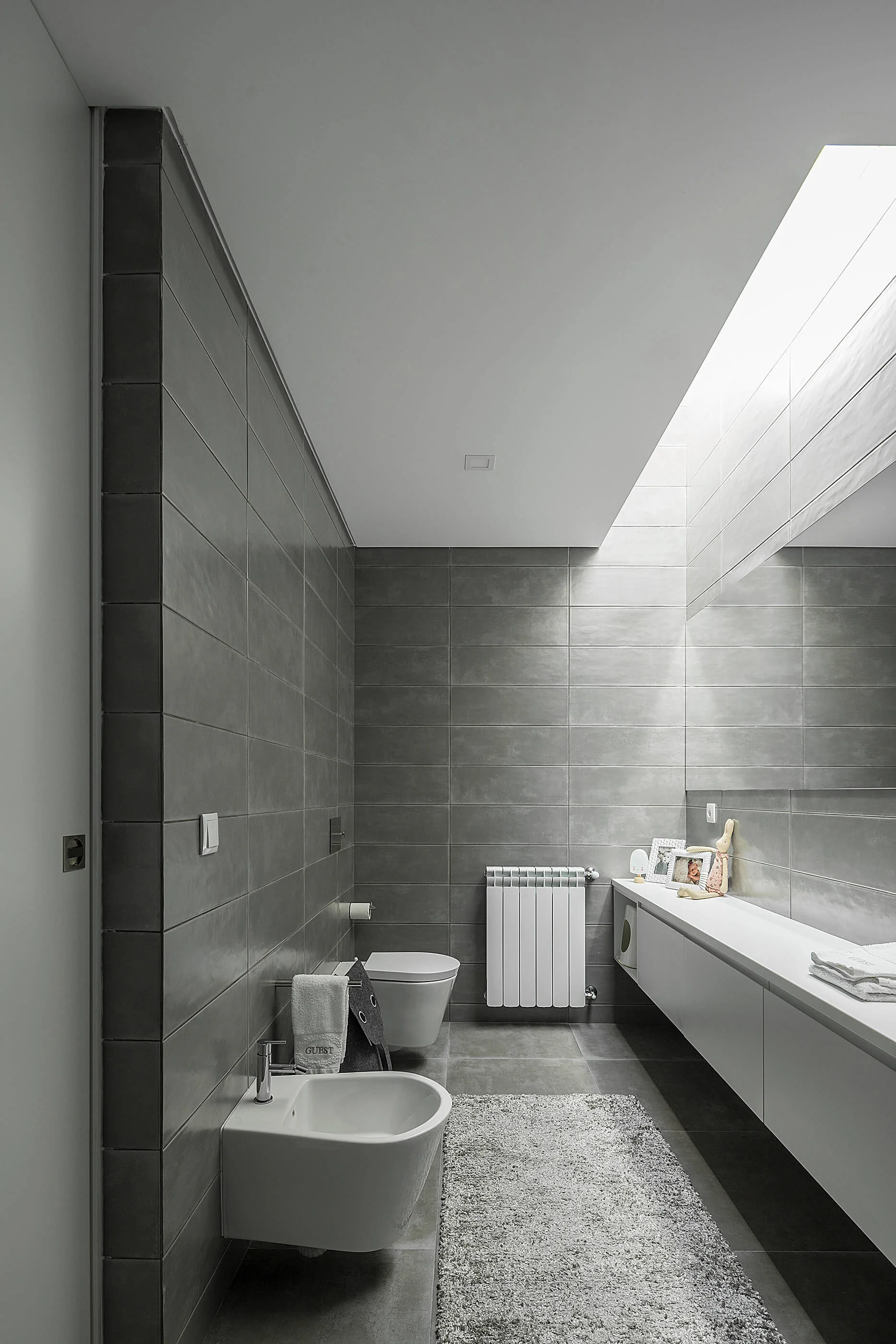 A minimalist contemporary bathroom with grey wall tiles, a white floating vanity, a bidet, a wall-hung toilet, and a long ceiling skylight.