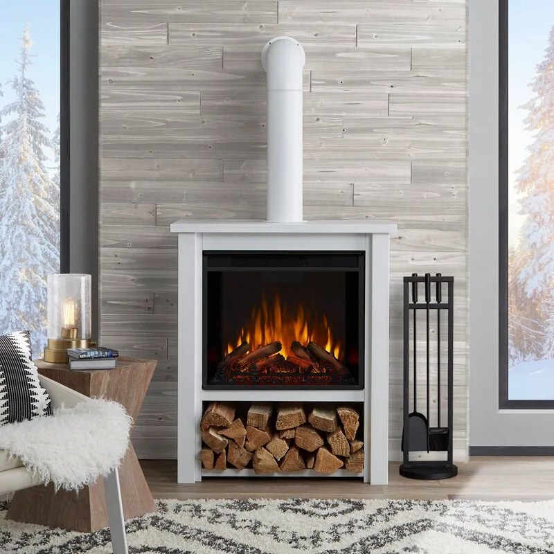 A freestanding white electric fireplace with stacked logs in its lower compartment sits against a grey wood-planked wall between two snowy windows.