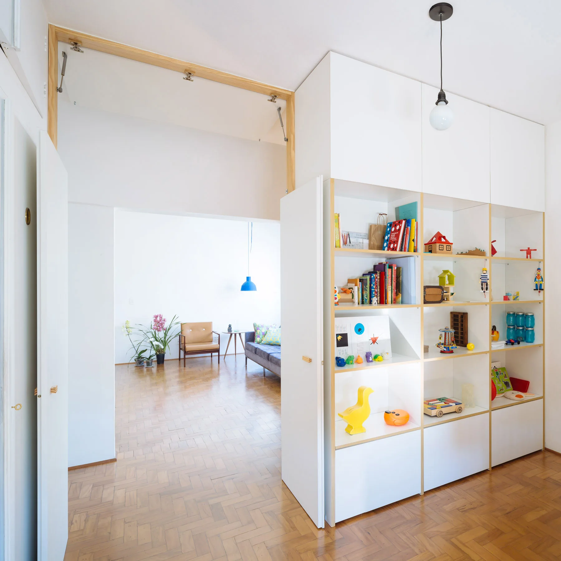 A custom white shelving unit filled with toys sits on herringbone parquet flooring next to a wide doorway opening into a living room.