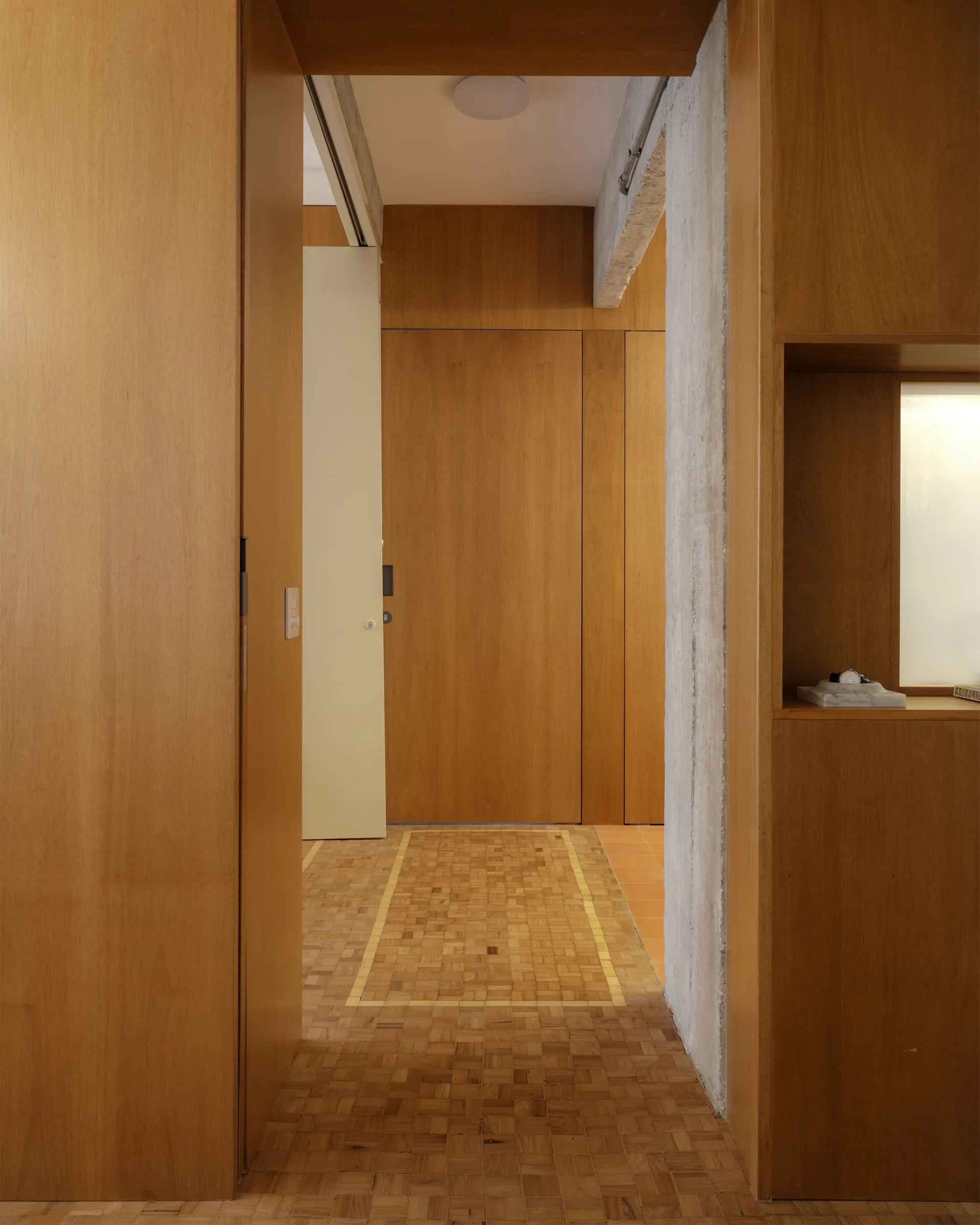 A modernist hallway featuring warm wood-paneled walls, geometric parquet flooring, an exposed concrete pillar, and a recessed wall niche.