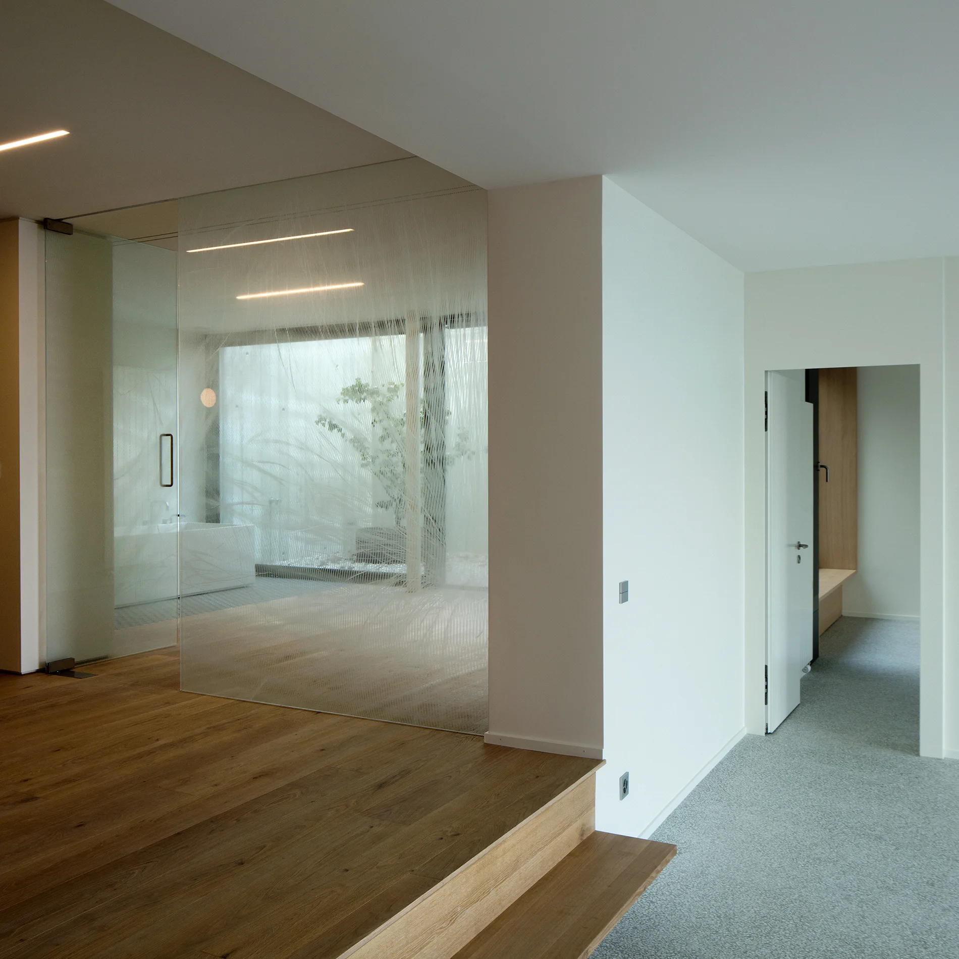 Oak wood steps lead to an etched glass partition wall screening a minimalist white bathroom with a bathtub.