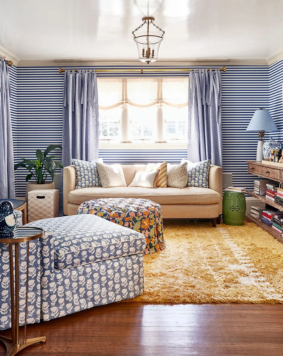 An eclectic living room featuring blue and white striped wallpaper, a beige sofa, a yellow shag rug, and patterned blue armchairs.
