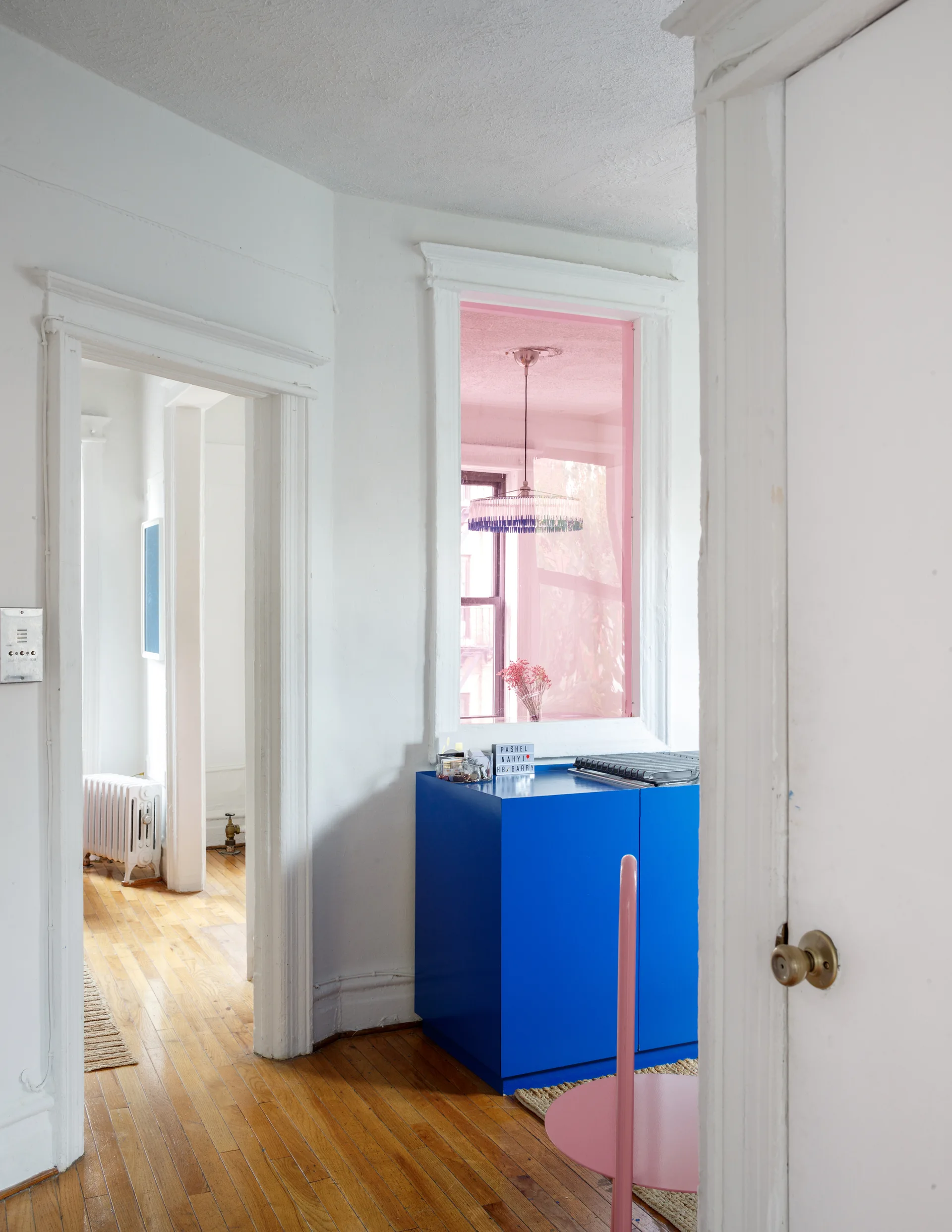 A white interior hallway with wood floors features a bright blue storage cabinet beneath a pink-tinted window and a pink chair.