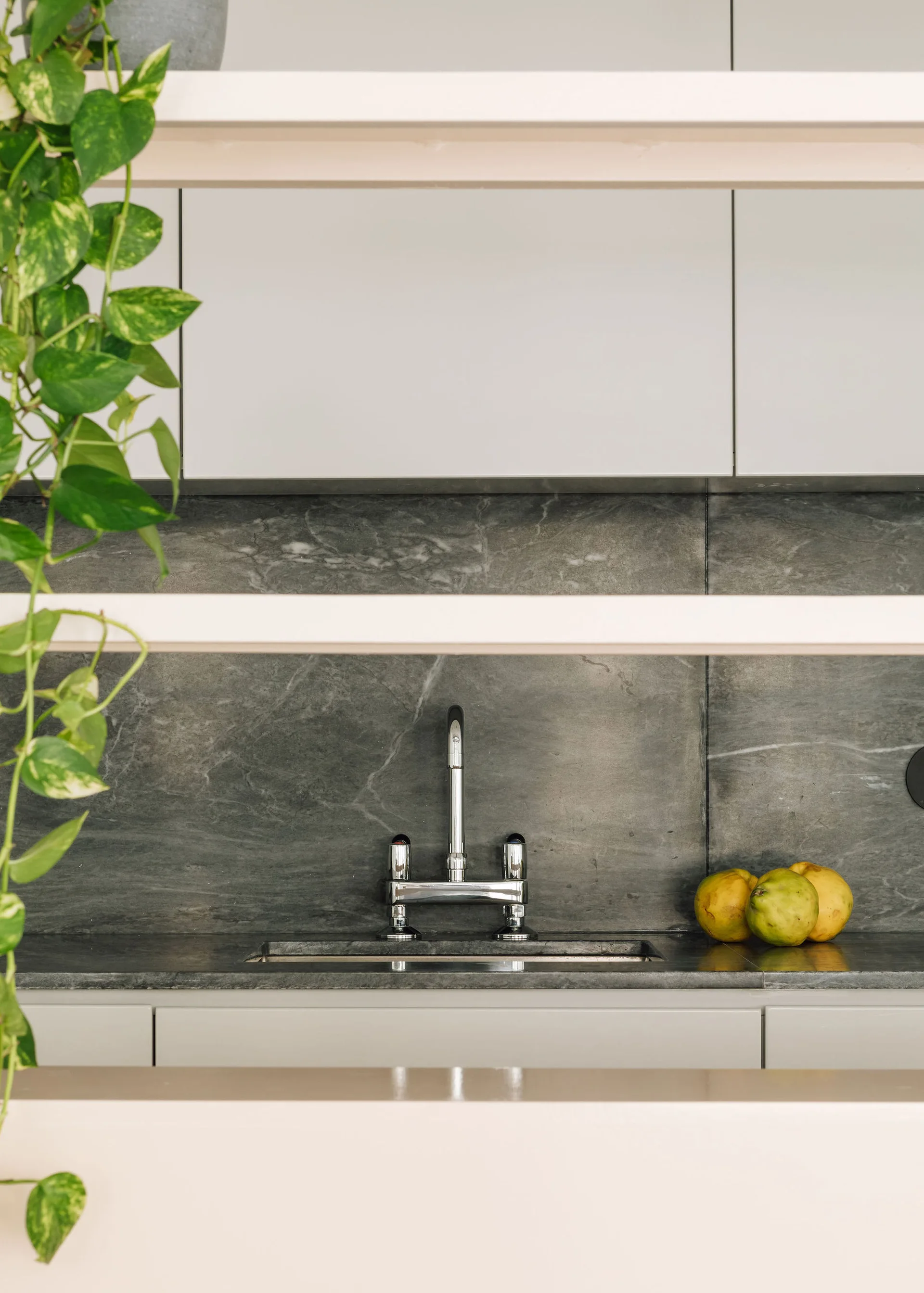 A kitchen sink with a chrome faucet set against a veined grey stone backsplash, framed by light open shelves with a trailing green plant.