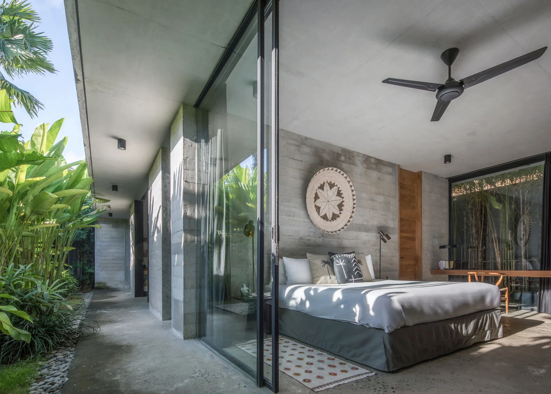 A modern bedroom features textured concrete walls, a bed with a woven wall hanging, and large glass sliding doors opening to a tropical garden pathway.