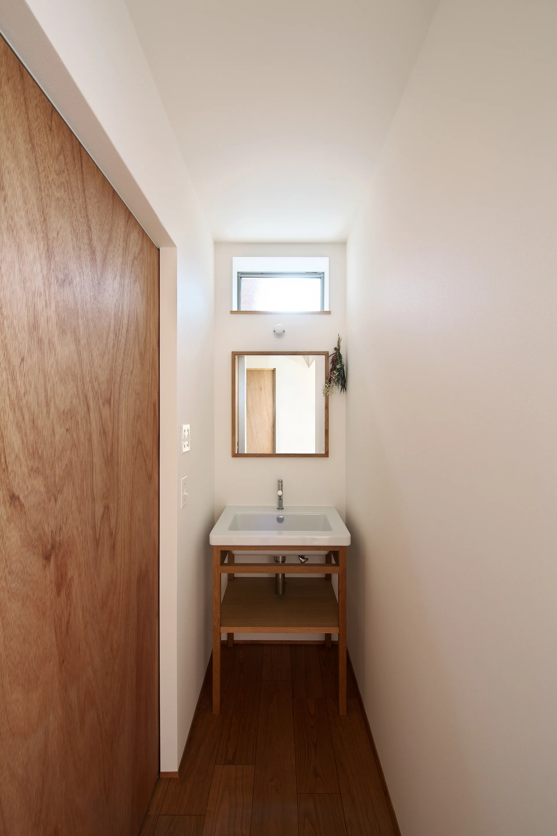 A narrow white powder room features an open-frame wooden vanity with a white ceramic sink, a square mirror, and a small high window.