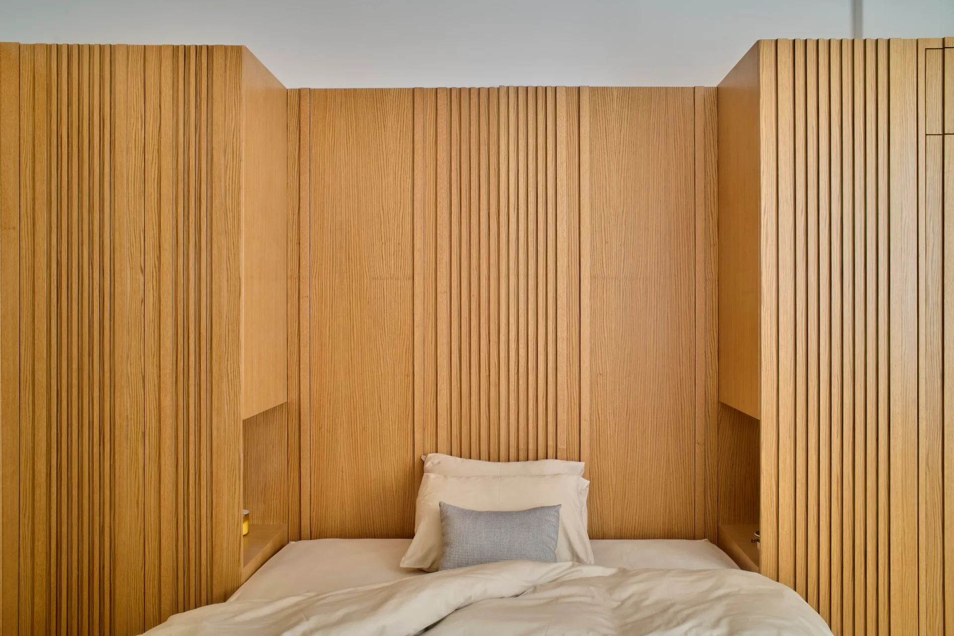 A minimalist bed is framed by a vertical slatted wood headboard and symmetrical built-in wardrobes with small recessed nightstand niches.