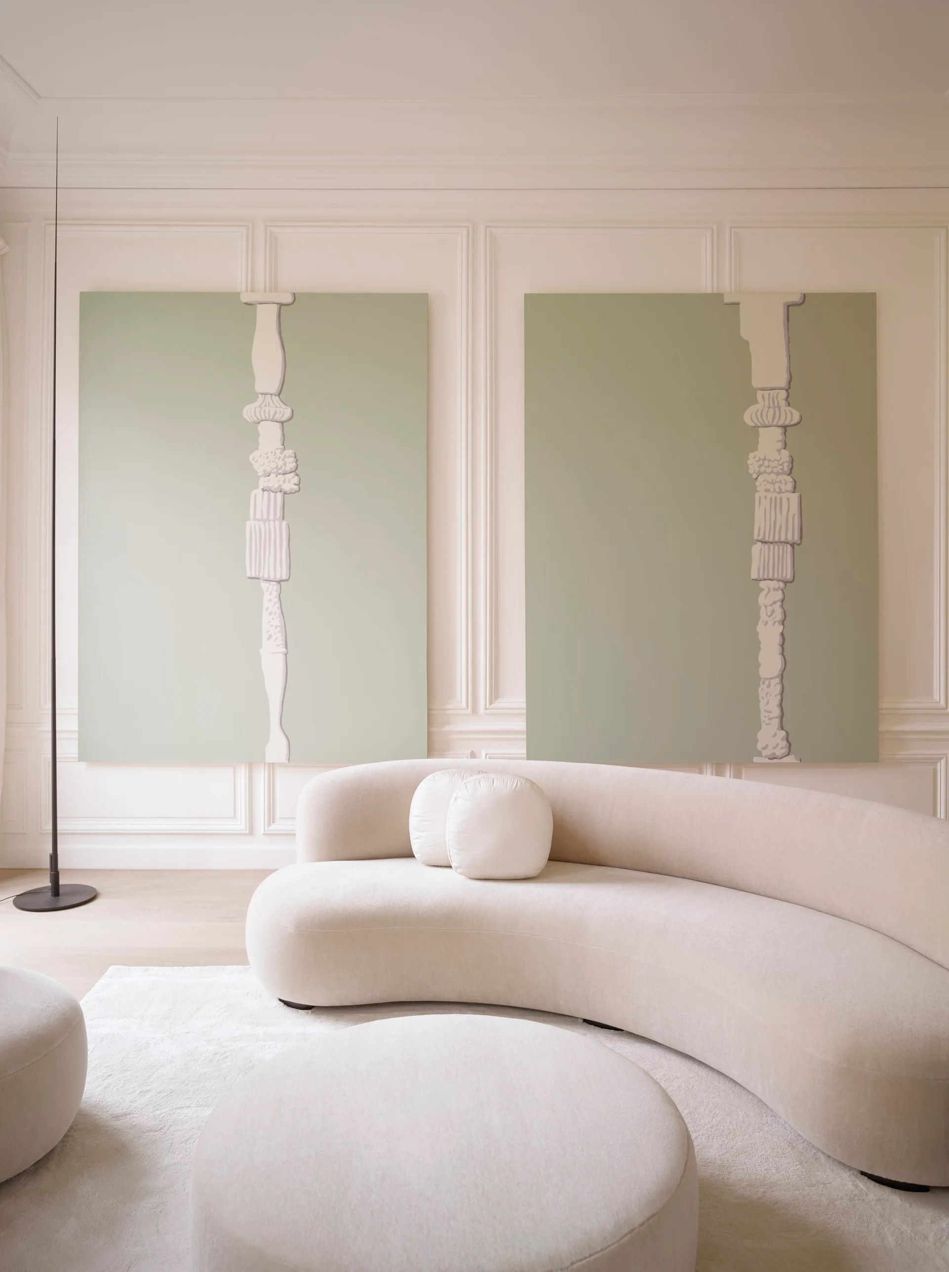 A cream curved sofa and round ottoman sit in front of an off-white wainscoted wall decorated with two large sage green abstract paintings.