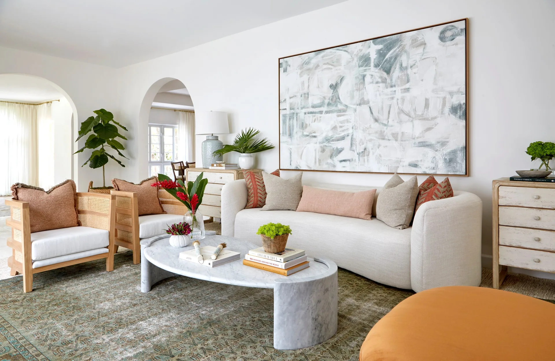 A bright living room features a cream curved sofa, an oval marble coffee table, wood-framed cane armchairs, and a large abstract painting.