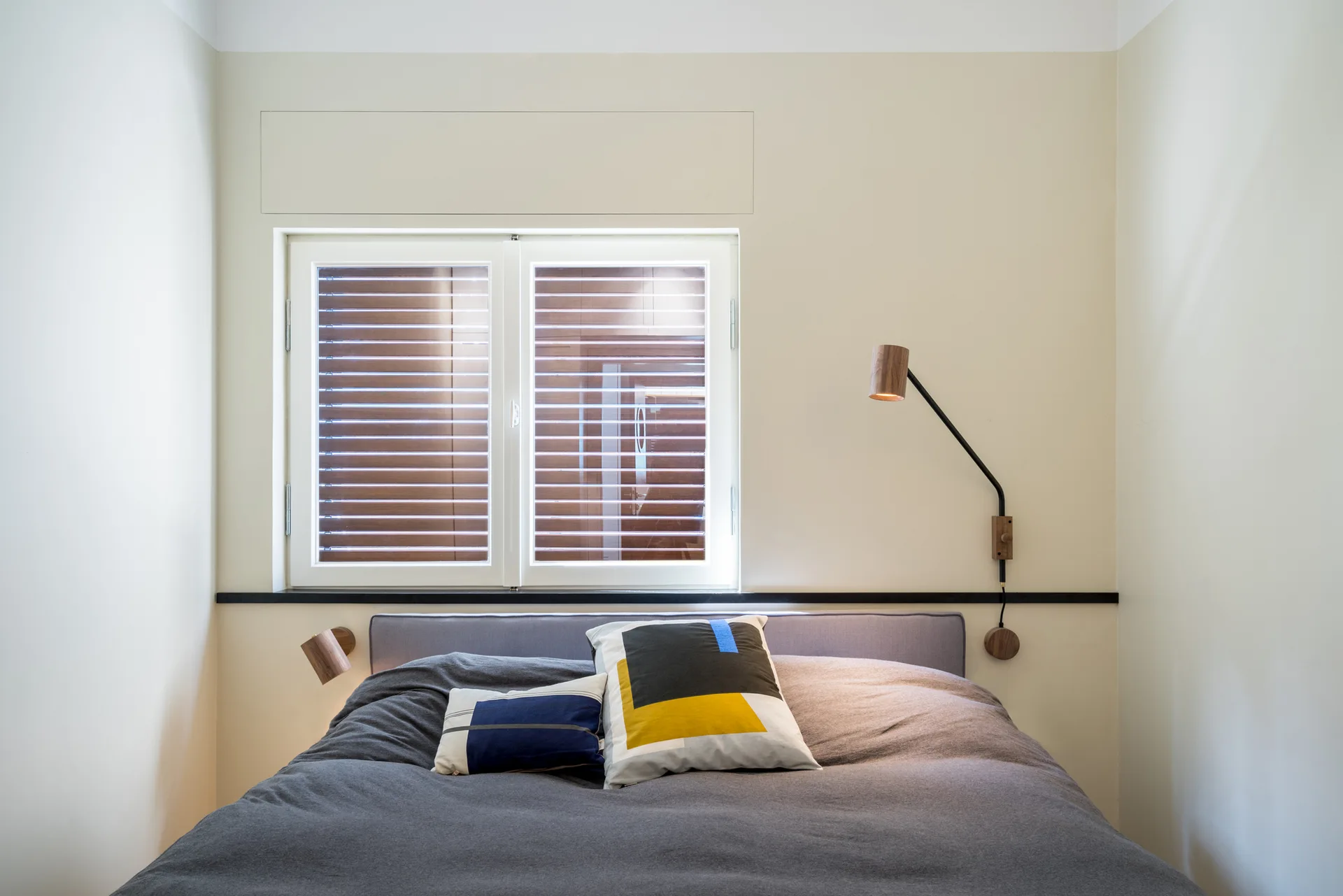 A grey upholstered bed with color-blocked pillows sits beneath a shuttered window, flanked by two wall-mounted wood and metal reading lights on a beige wall.