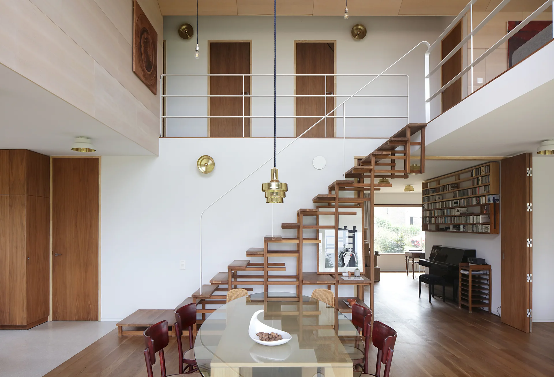 An open wooden staircase connects a mezzanine level to a dining area featuring a glass top table with wooden chairs.
