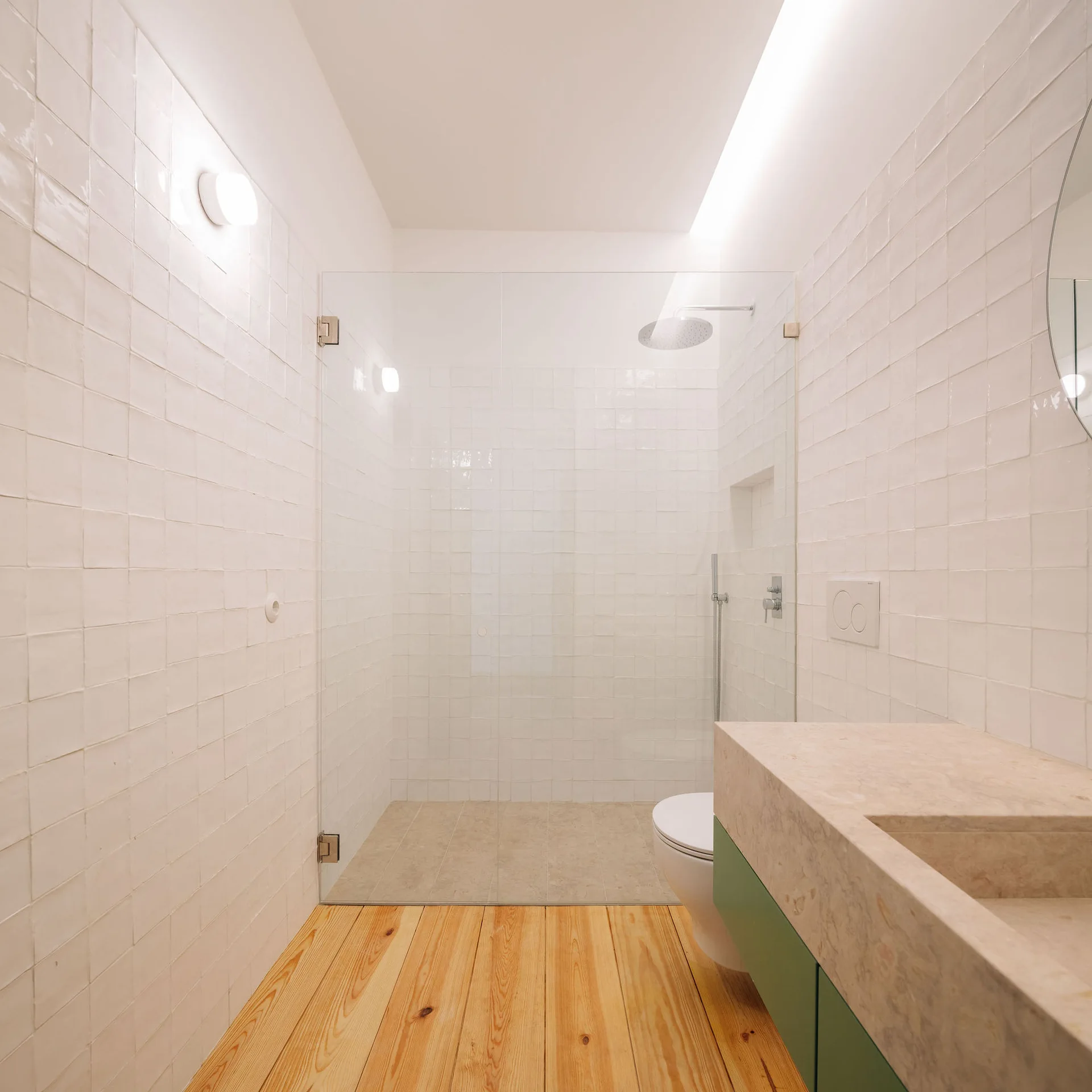 A minimalist bathroom with glossy white square tiles, a light wood plank floor, a glass shower partition, and a green vanity with a beige stone countertop.