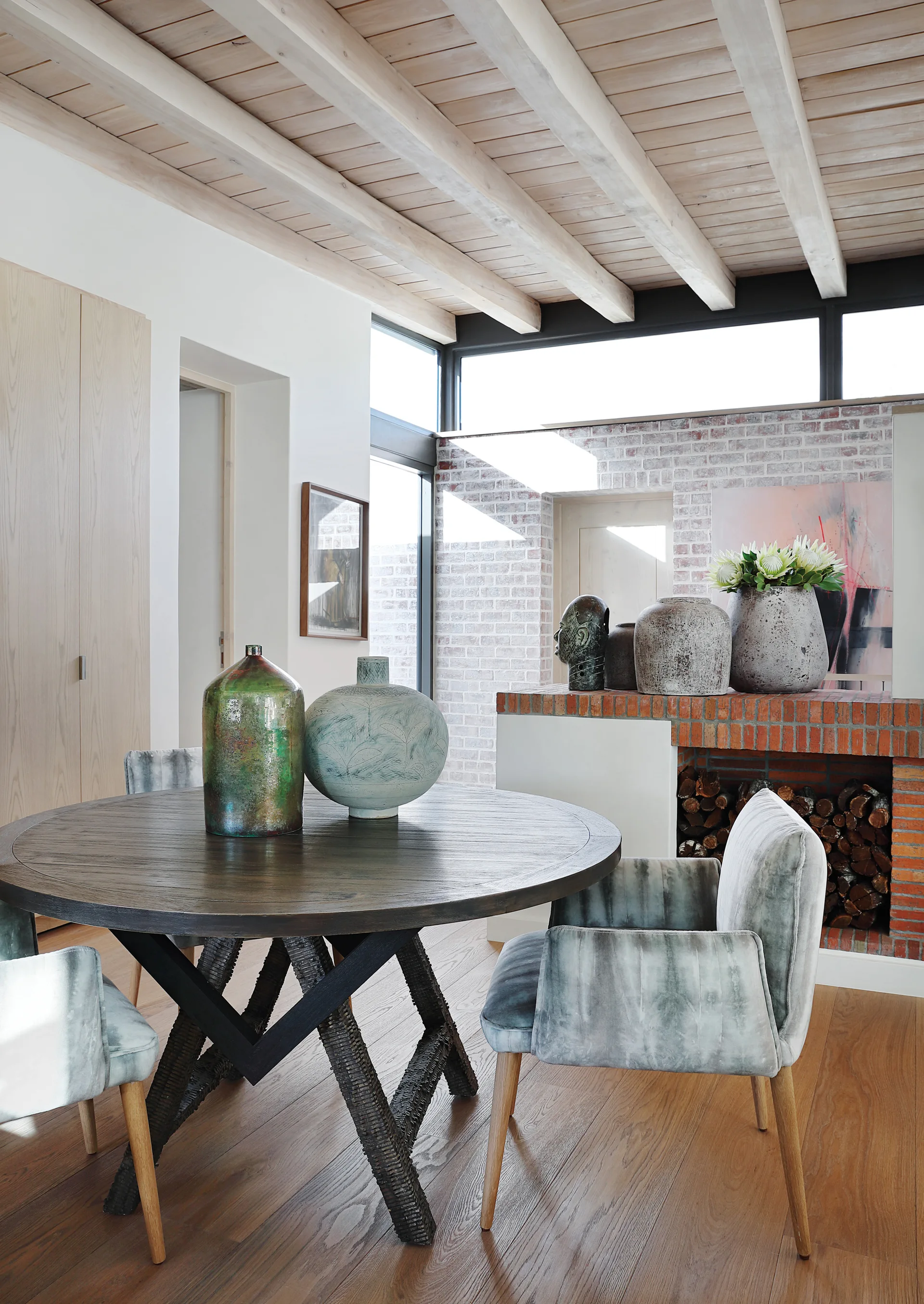 A round dark wood dining table surrounded by mottled grey-green chairs sits under a light wood beamed ceiling next to an exposed brick fireplace.