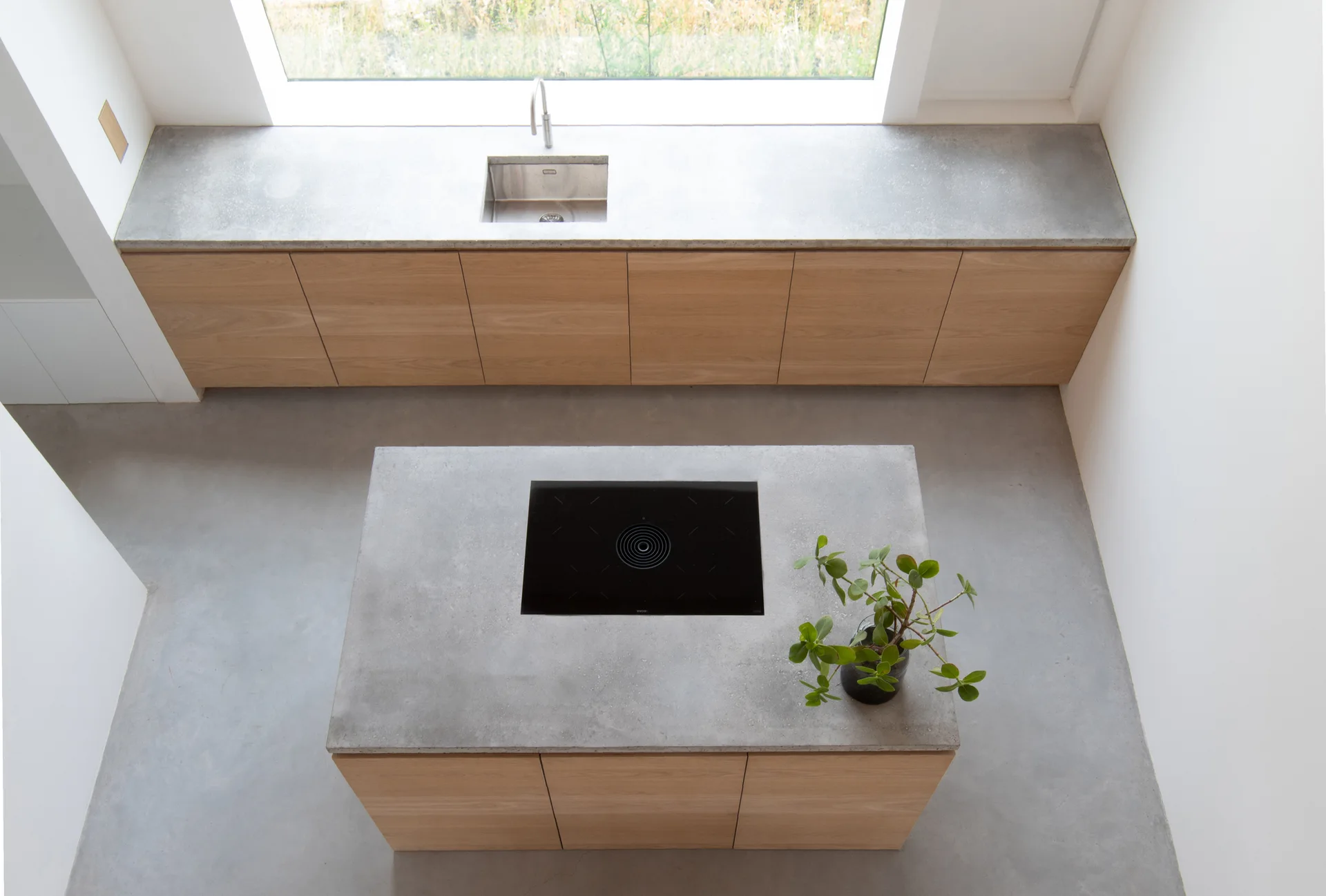 Overhead view of a modern kitchen with light wood cabinets, gray concrete-like countertops, a black cooktop on the island, and a stainless steel sink.