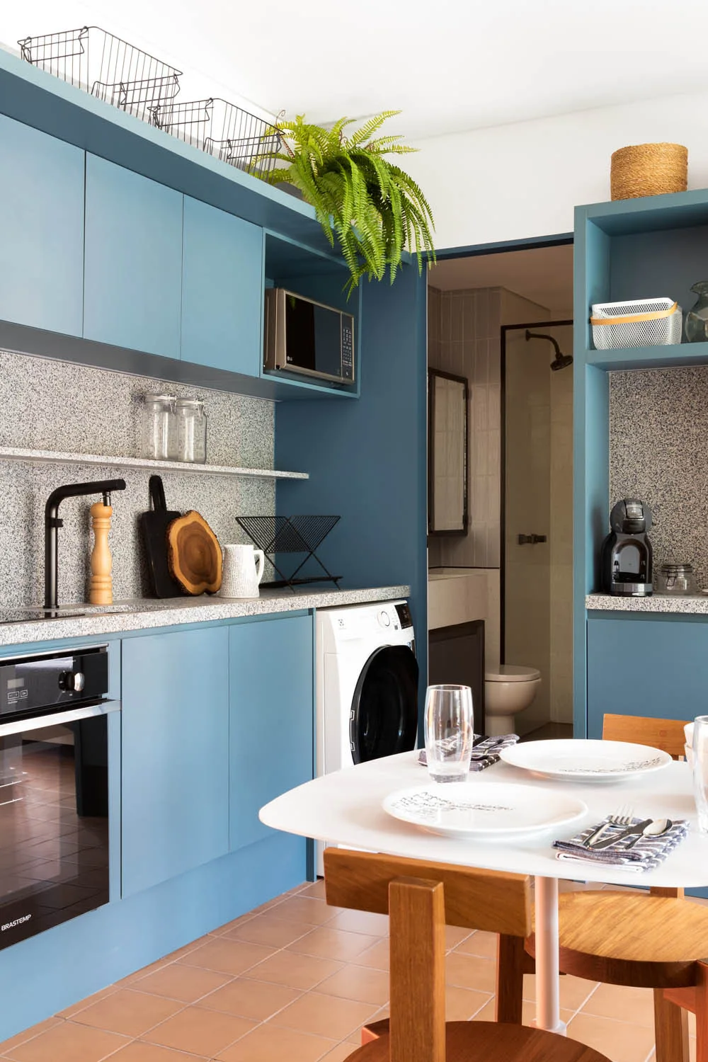 A compact kitchen features matte blue cabinets, a speckled grey countertop, a built-in washing machine, and a small dining table with wooden chairs.