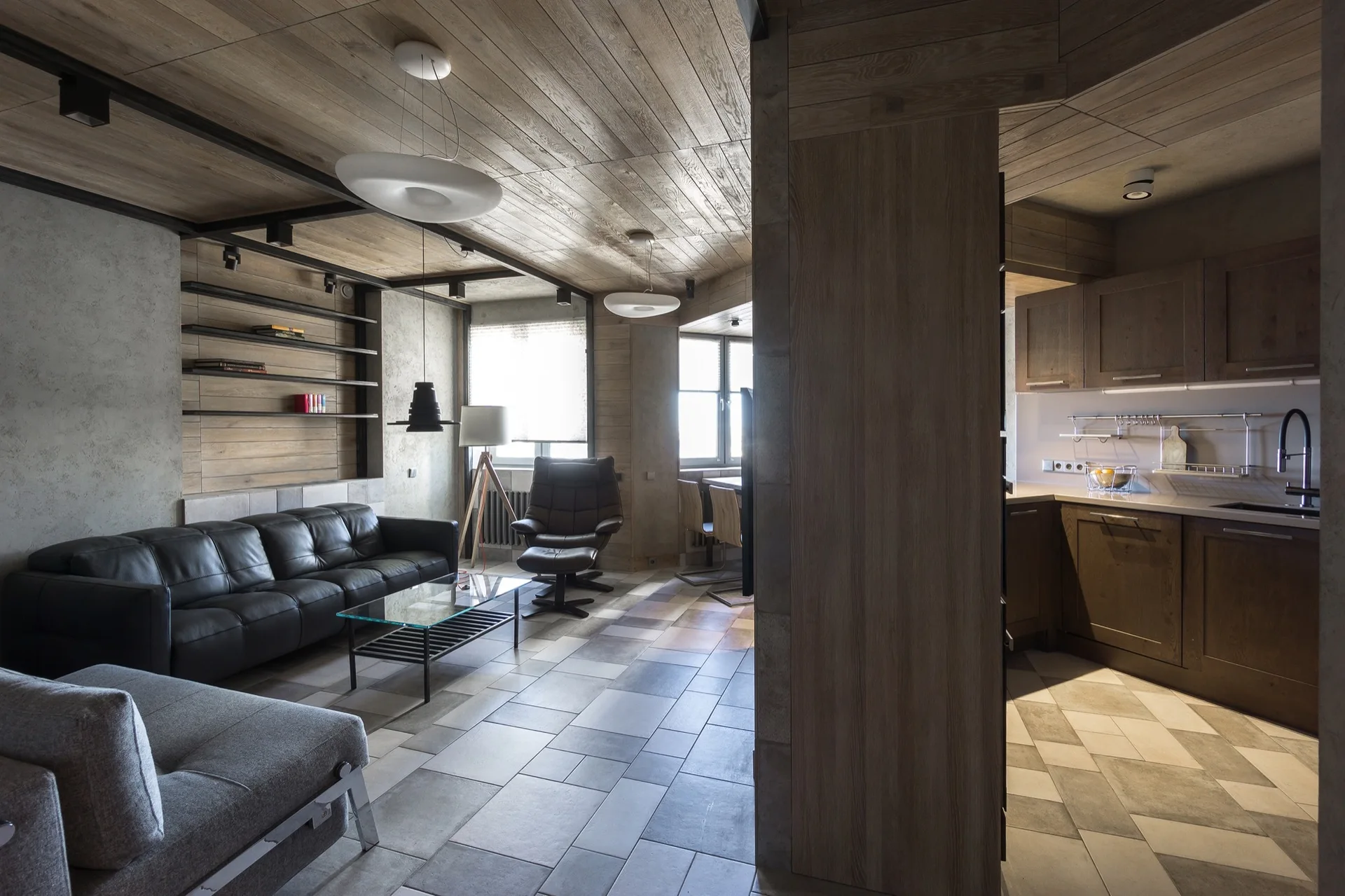 A contemporary open-concept living room featuring a black leather sofa, wood paneled ceiling, gray tile floor, and an adjacent kitchen with wood cabinetry.