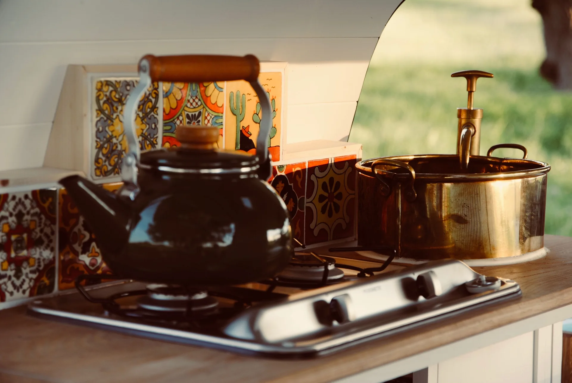 A compact kitchen counter features a black kettle on a gas stove next to a brass basin sink and a backdrop of vibrant, patterned ceramic tiles.