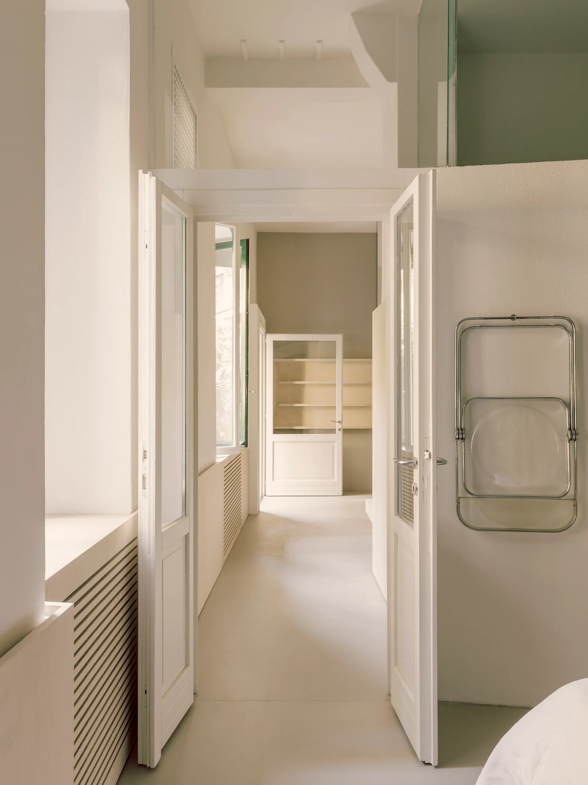 An open double door leads through a bright minimalist hallway featuring a slatted radiator cover and a folding chair hanging on the wall.