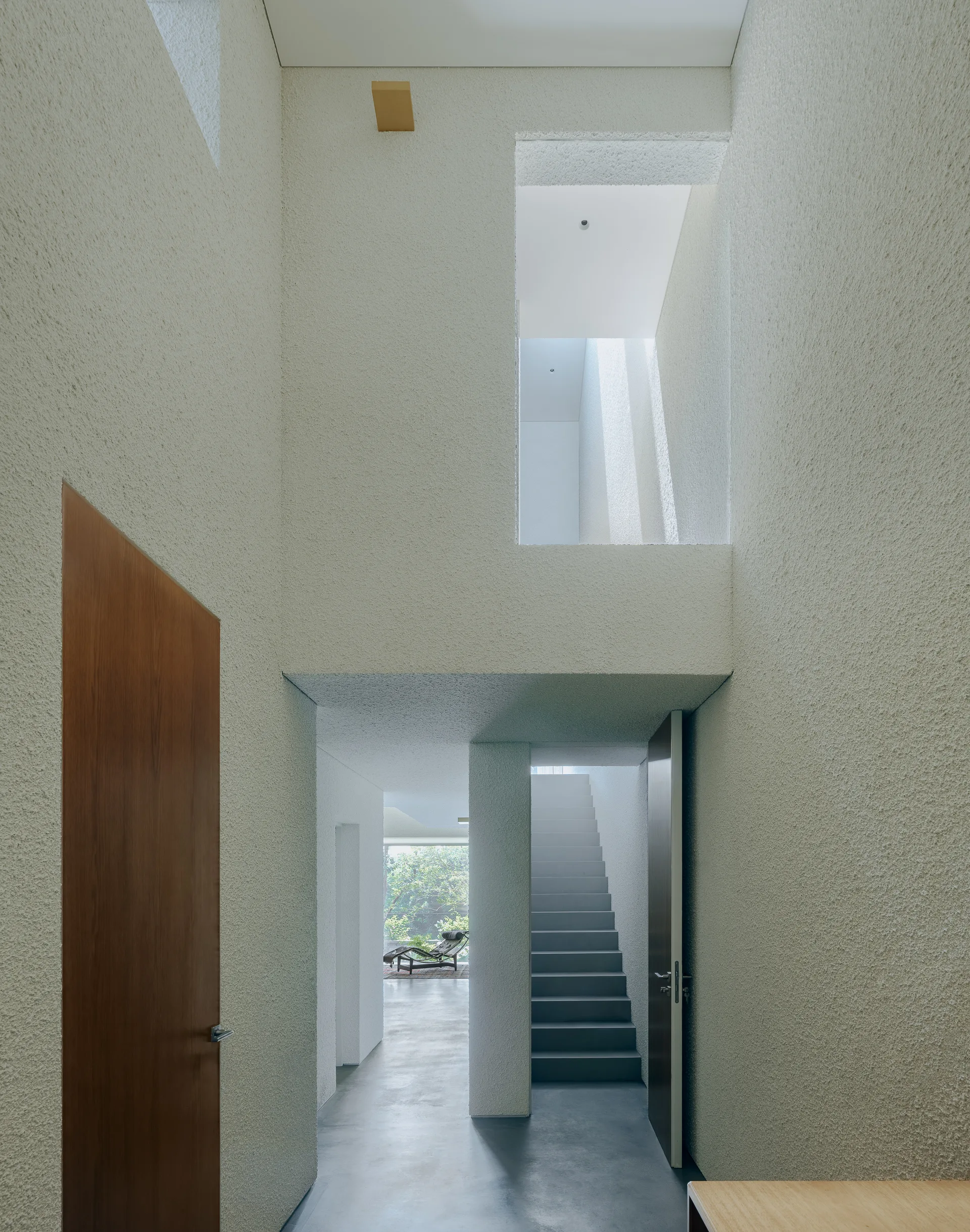 A minimalist residential entryway features heavily textured plaster walls, a smooth concrete floor, a straight staircase, and tall wood doors.