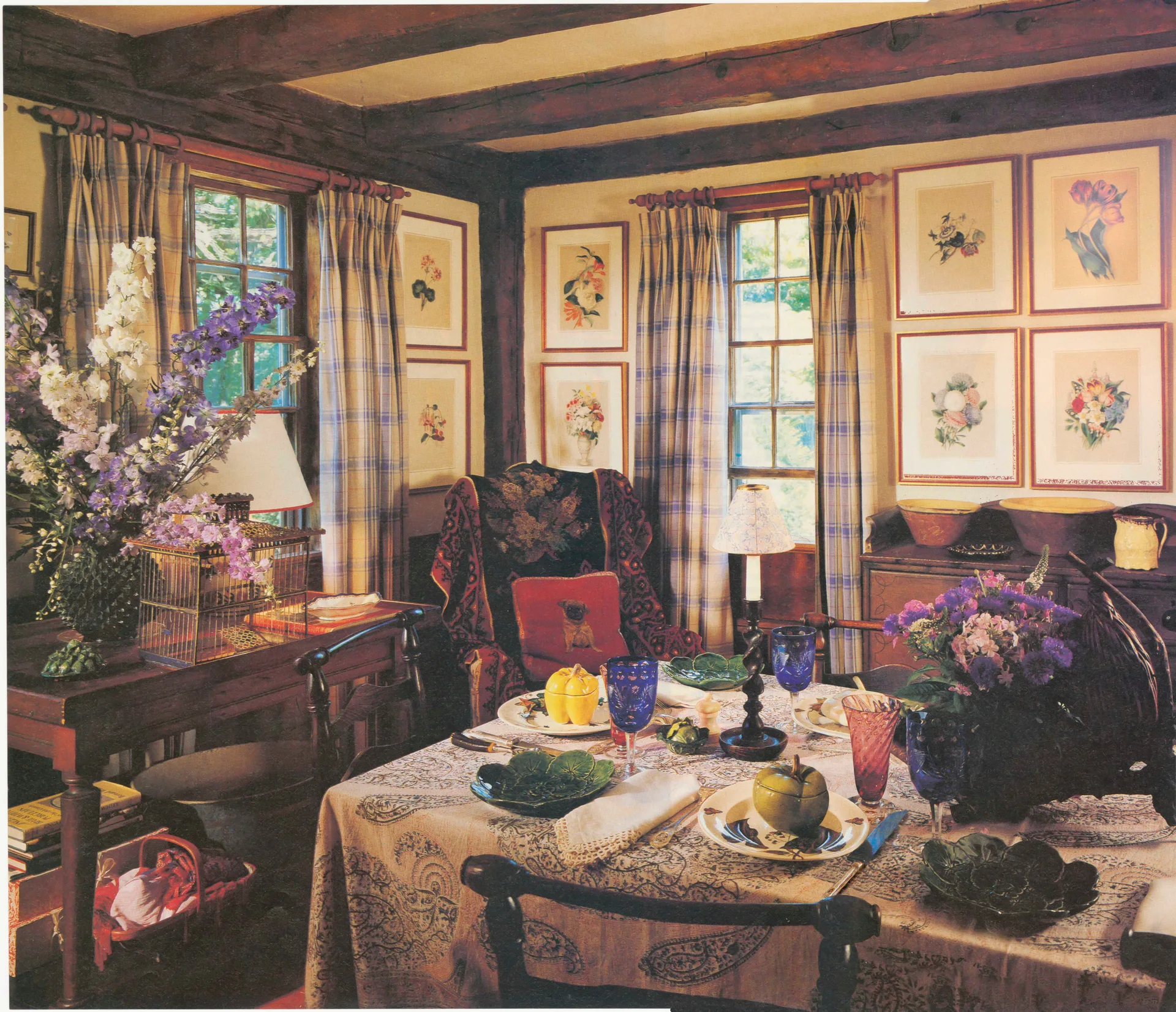 A rustic dining room features a table set with paisley print linens, surrounded by wood ceiling beams, plaid curtains, and botanical art prints.