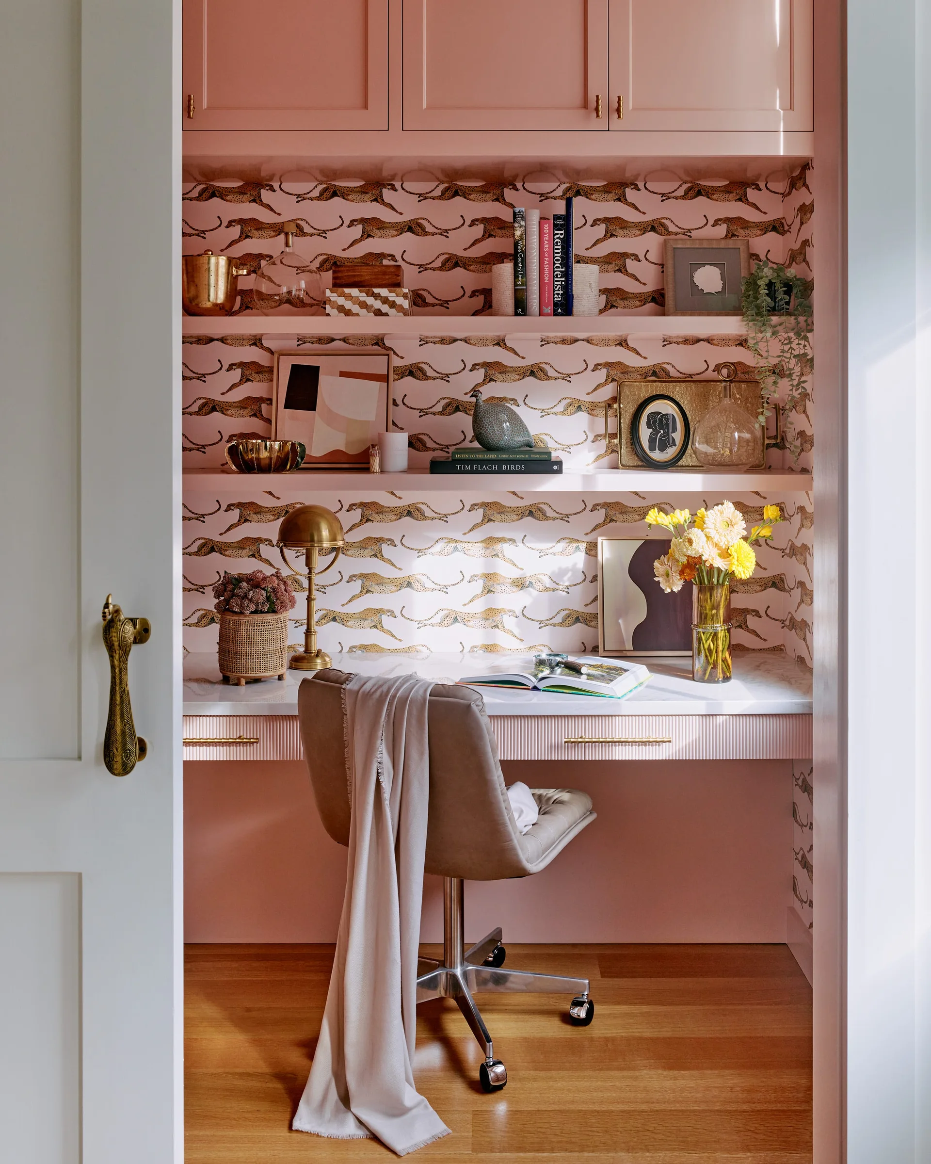 A compact home office nook features a pink built-in desk, running leopard wallpaper, matching shelves, and a gray swivel chair.