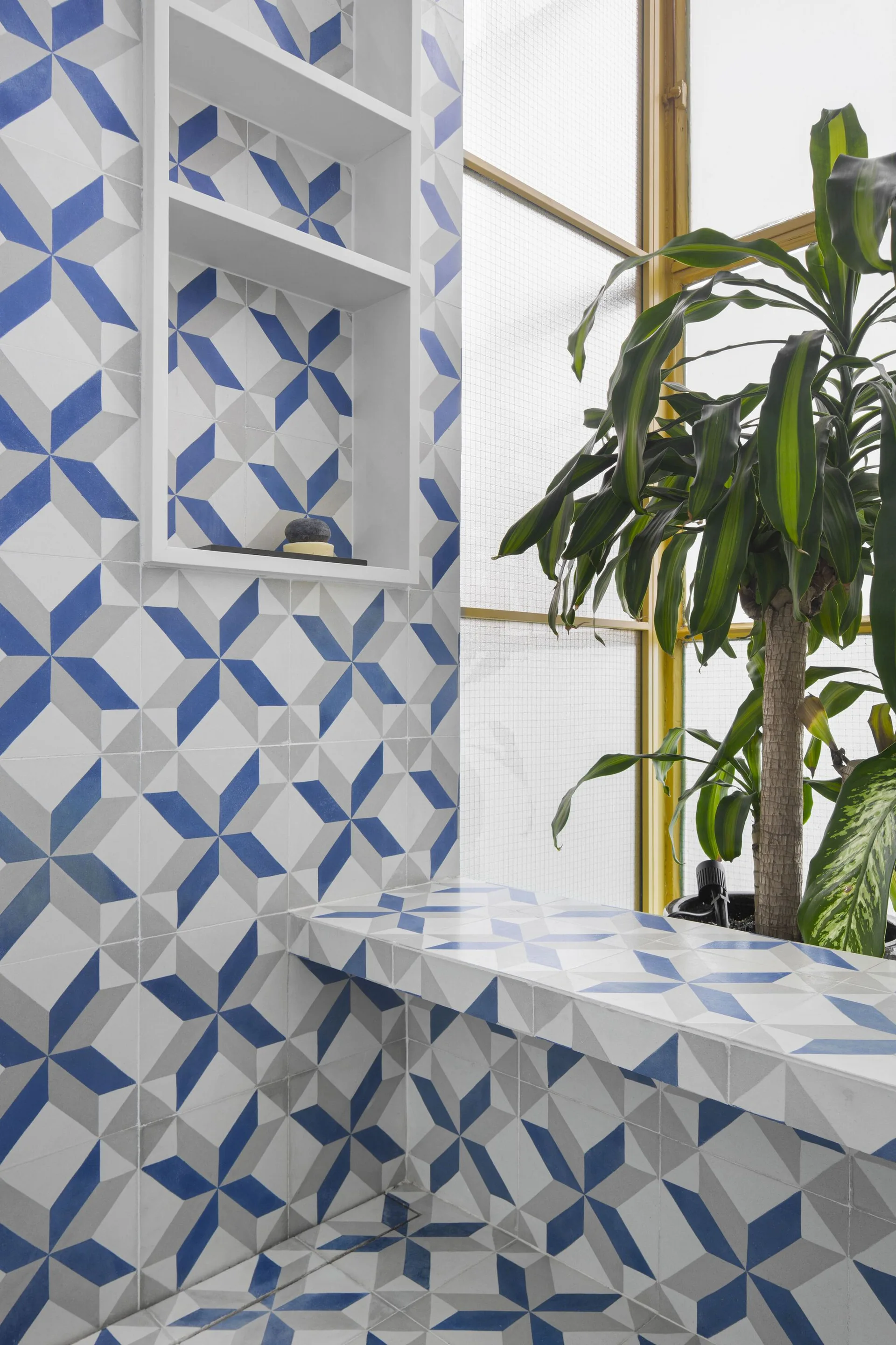 Bathroom corner featuring blue and grey geometric star tiles on the walls and bench, a recessed white niche, and a green potted plant by a window.