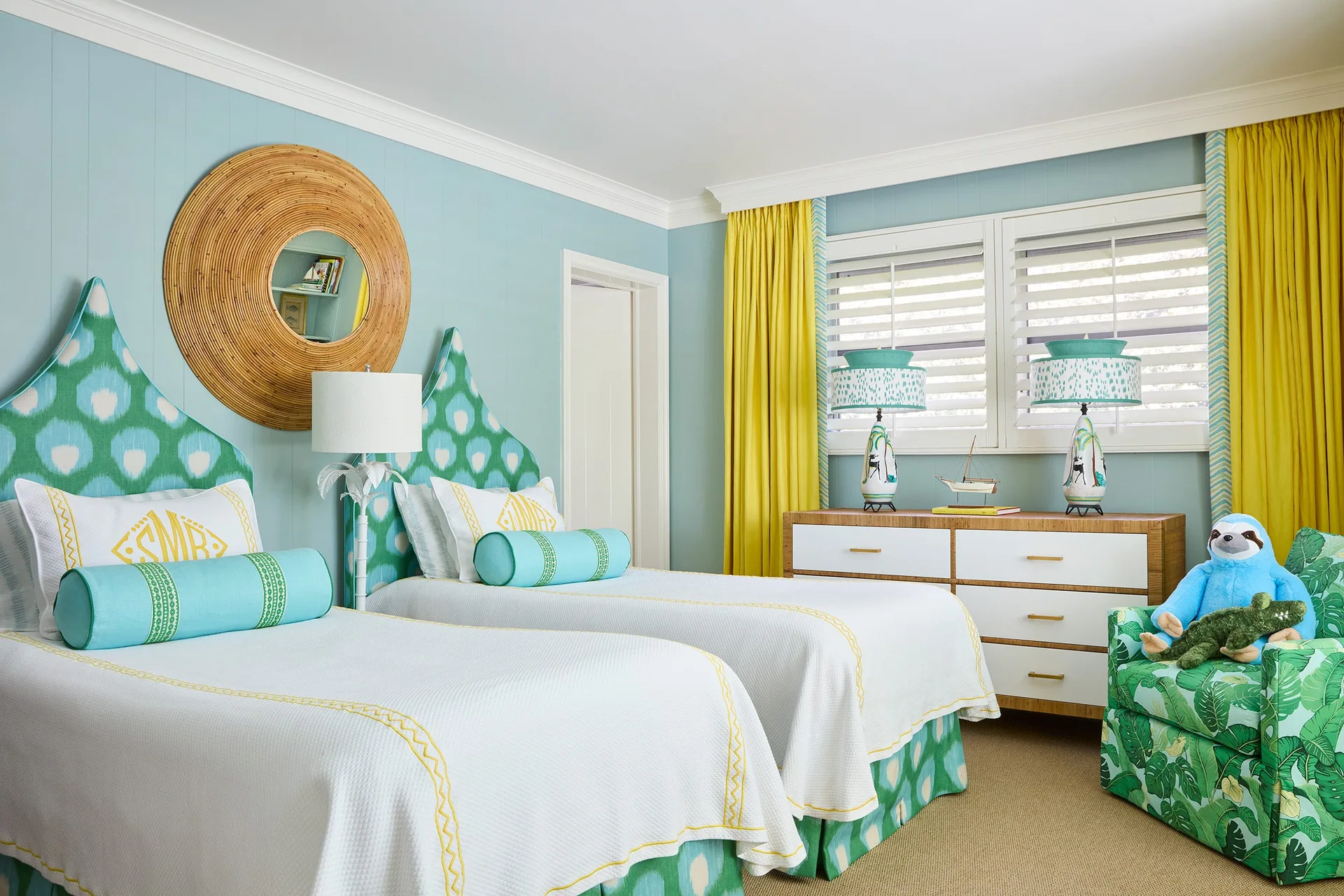 Two twin beds with green ikat headboards stand against light blue paneled walls in a bedroom with yellow curtains and a white dresser.