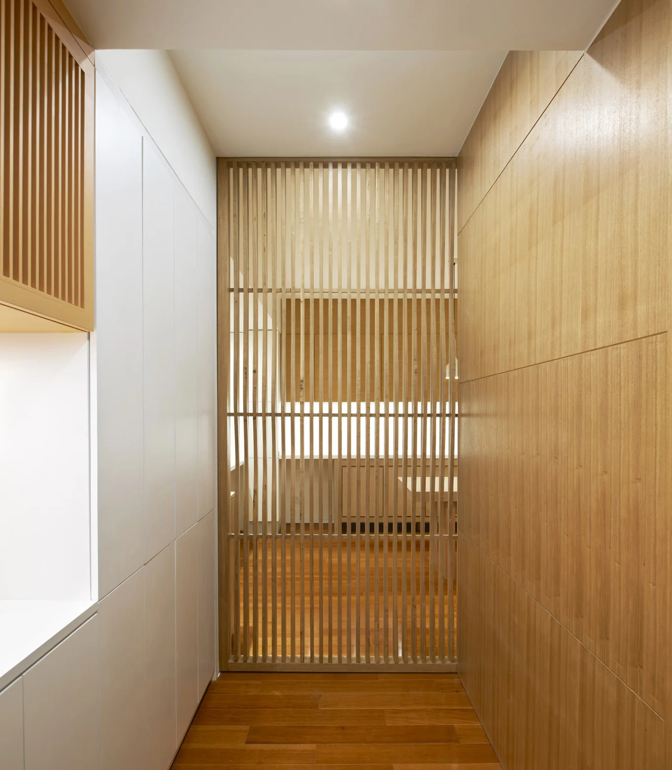 A narrow hallway features a vertical wood slatted room divider at the end, flanked by white built-in cabinets and warm wood wall paneling.