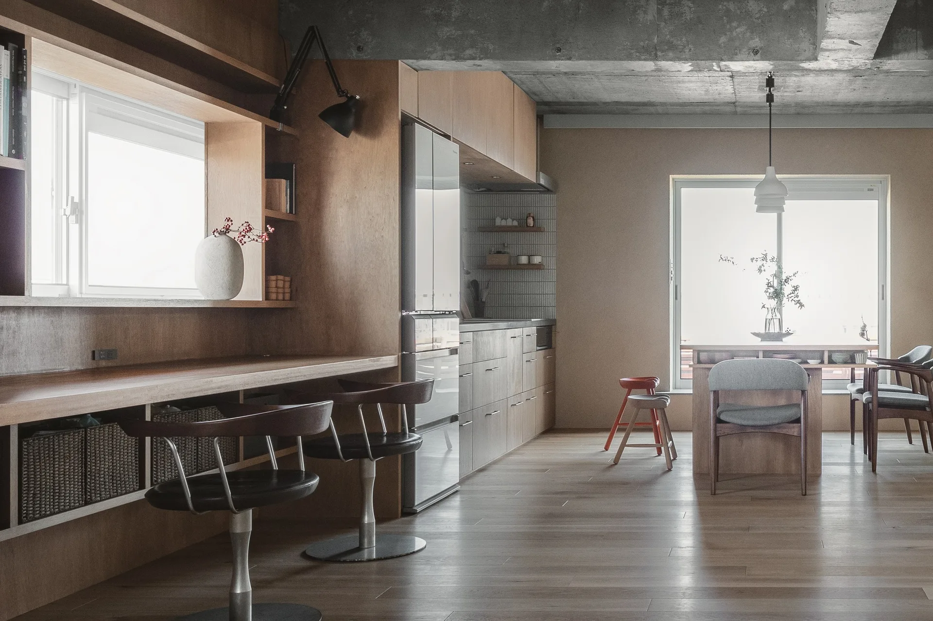 A modern kitchen and dining space with warm oak cabinetry, a built-in wooden desk, swivel bar stools, and an exposed raw concrete ceiling.