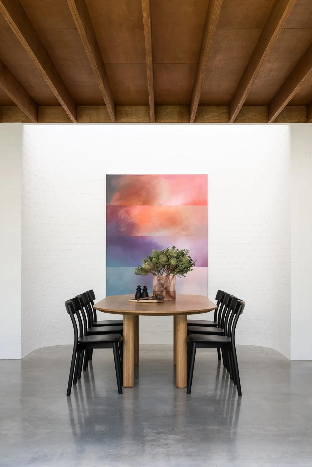 A wooden dining table with black chairs is set in a room with a curved white brick wall, a colorful abstract painting, and exposed wood ceiling beams.